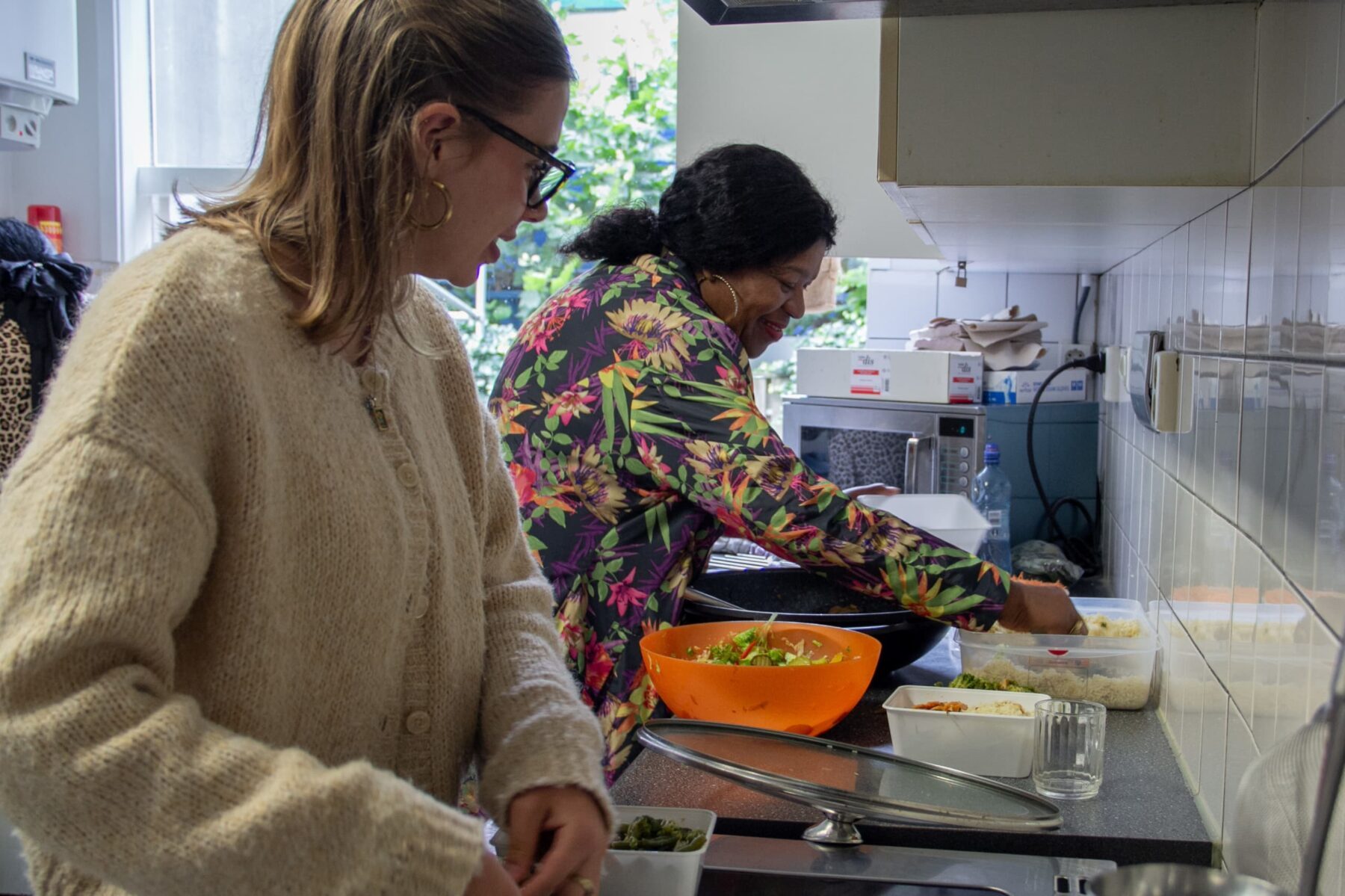 Zwei Freiwillige in kleiner Küche bereiten Essen vor; links beiger Wollpullover, rechts bunte Blumenjacke, orange Schüssel.