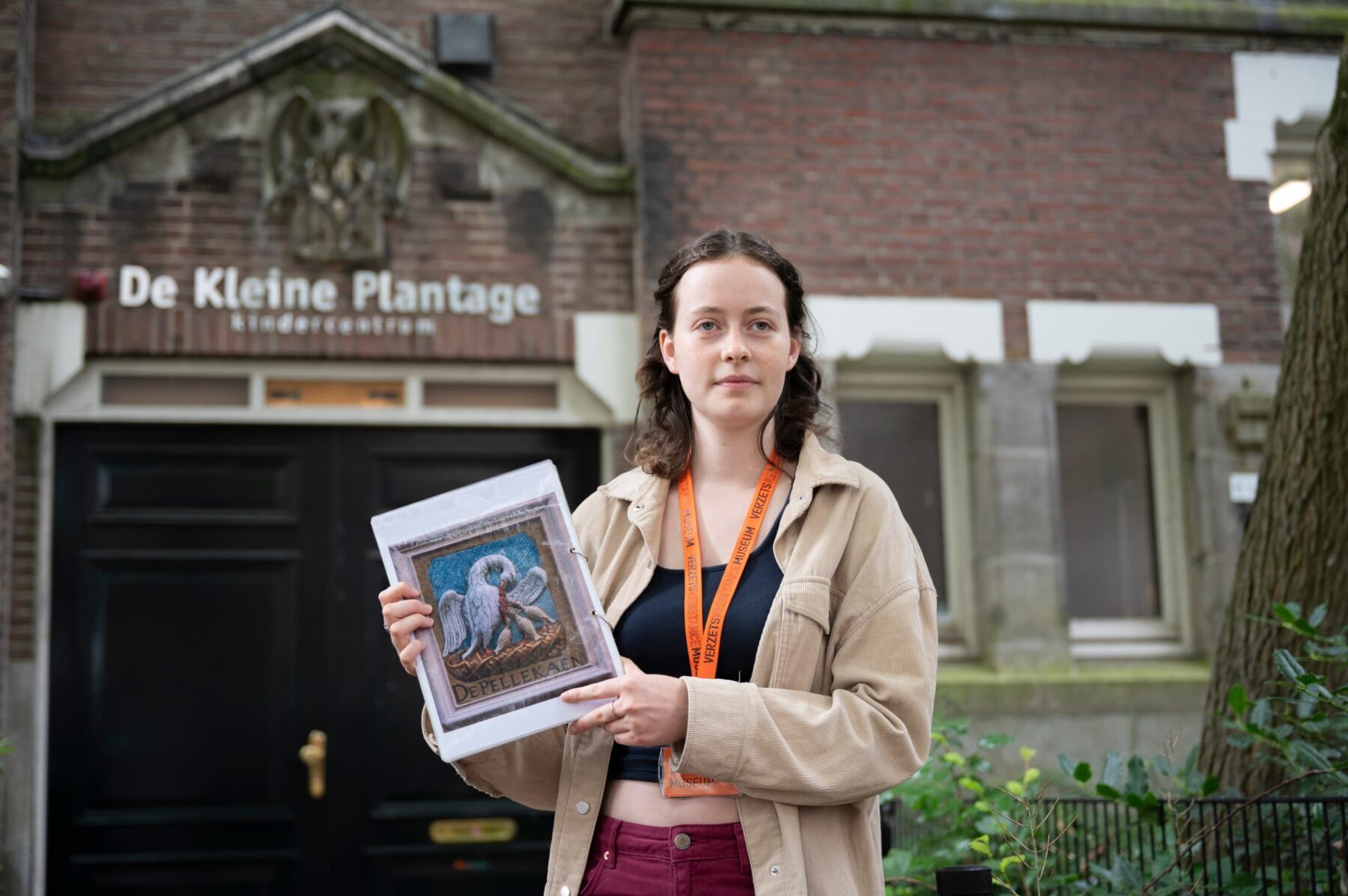 Freiwillige des Verzetsmuseum vor Eingang 'De Kleine Plantage', hält gerahmtes Mosaik mit Vogel, orange Lanyard, beige Jacke.