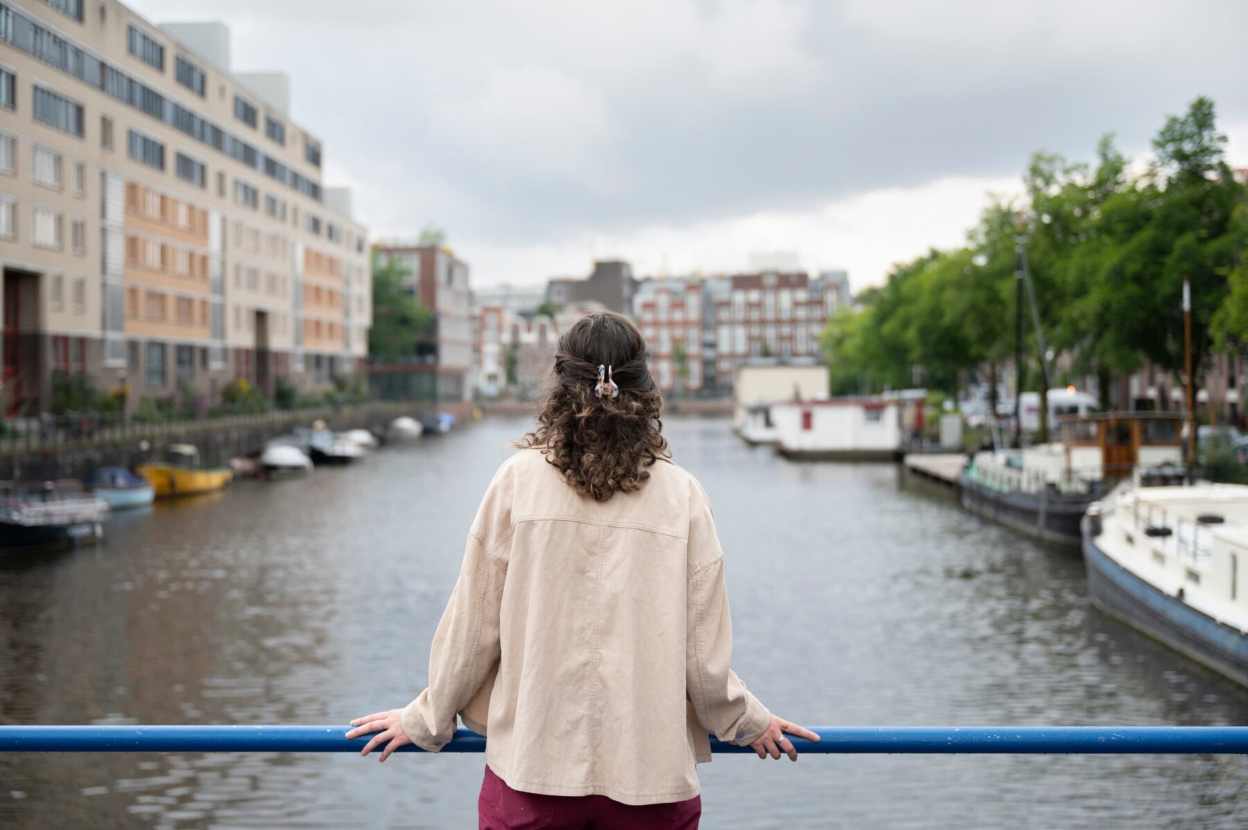 Freiwillige von hinten an blauem Geländer, beige Jacke, lockiges Haar, Blick auf Amsterdamer Kanal mit Hausbooten und Häusern.