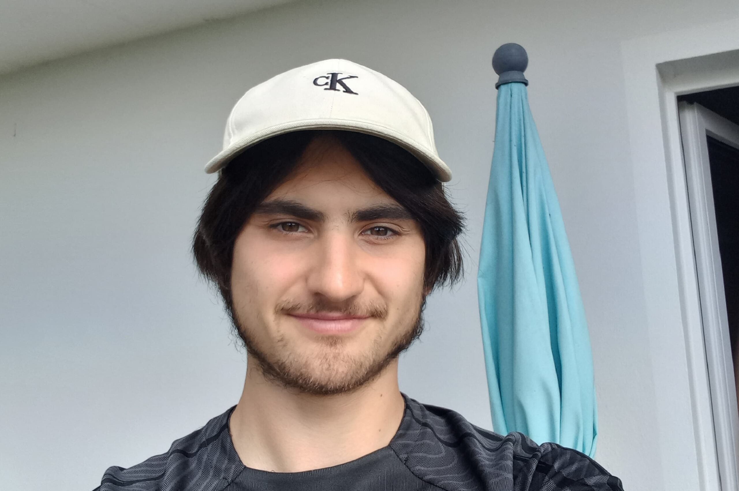 Mann mit weißer cK-Kappe und schwarzem Shirt, Gesicht verpixelt, hellblauer Sonnenschirm neben ihm.