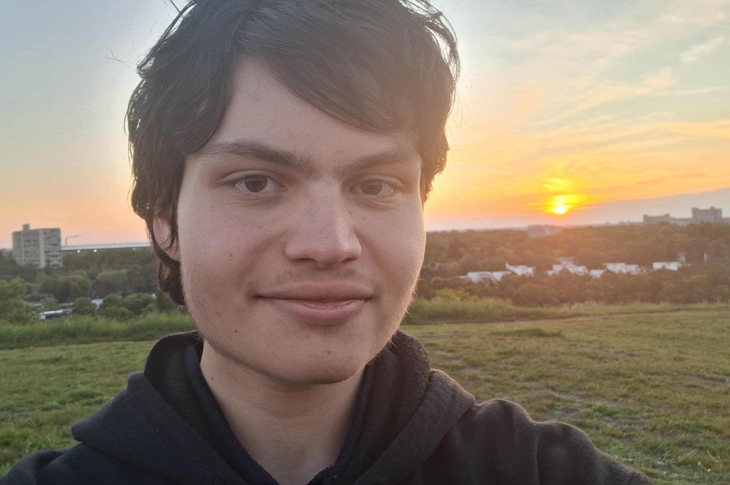 Person im dunklen Hoodie (Gesicht unkenntlich) beim Selfie auf grasigem Hügel, Sonnenuntergang über Stadt und grünen Feldern