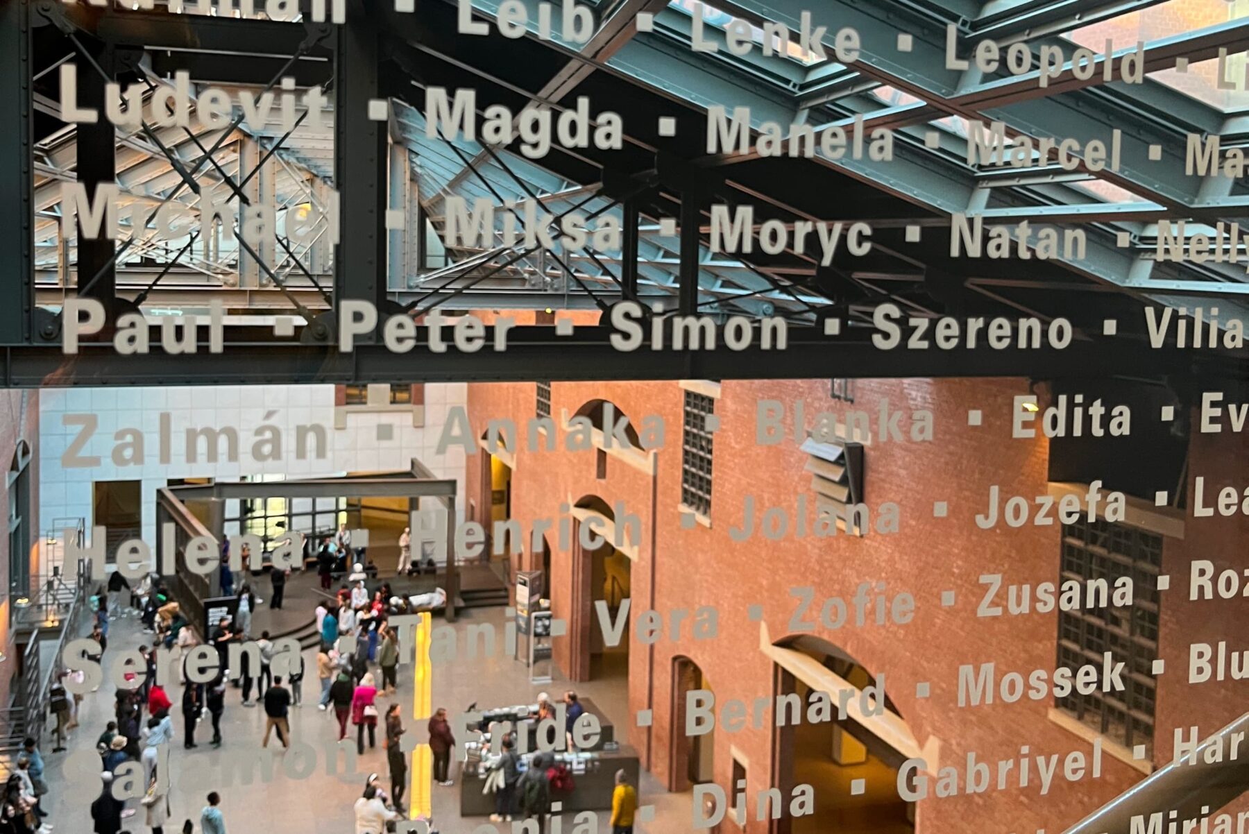 Glaswand mit aufgedruckten Namen, dahinter Blick ins Ausstellungsfoyer des USHMM mit Besuchern und rotem Backstein.