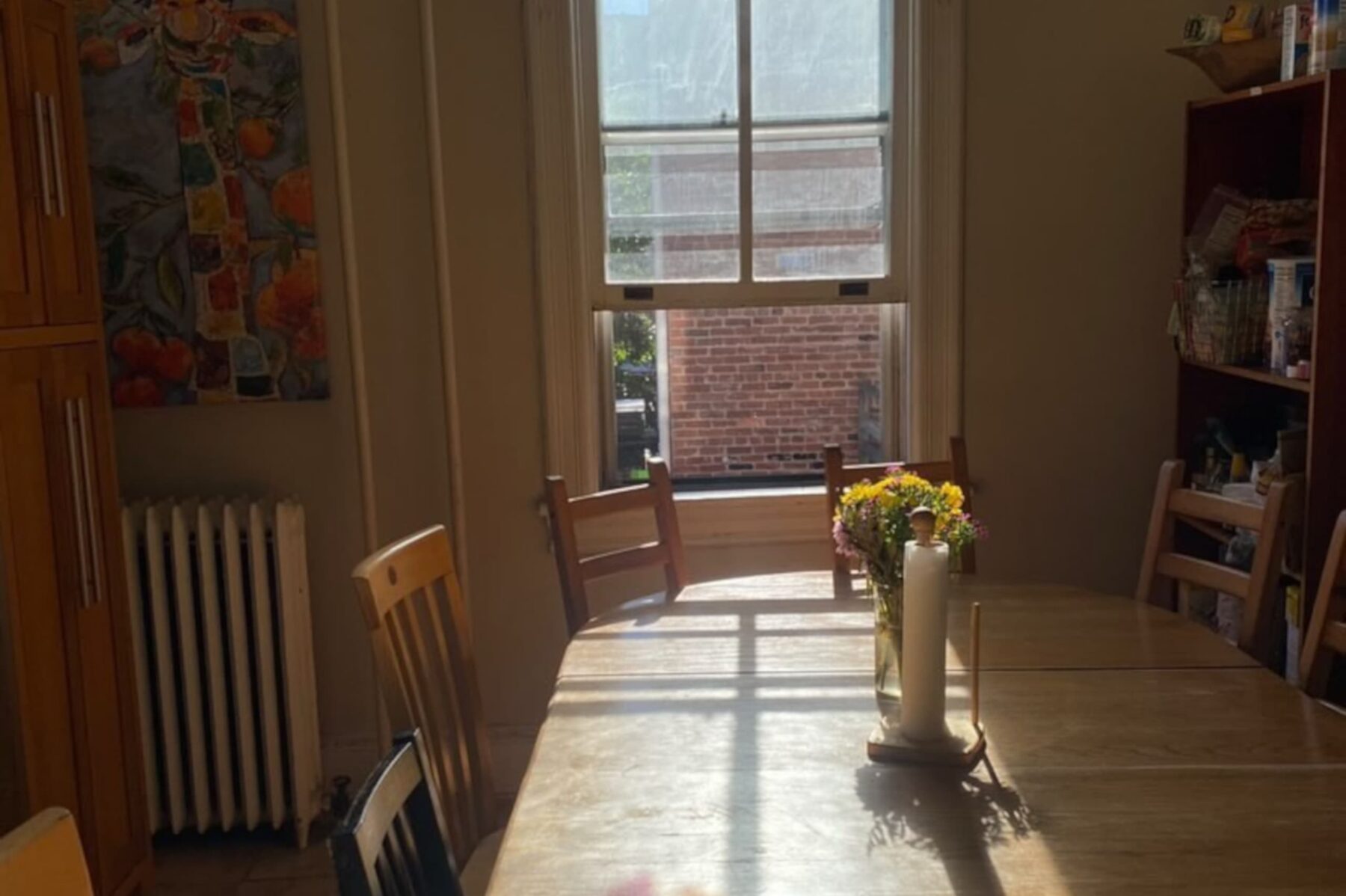 Heller Esstisch im Brethren House mit Holzstühlen, Blumenvase und Küchenrollenhalter, Sonnenlicht durch Fenster.
