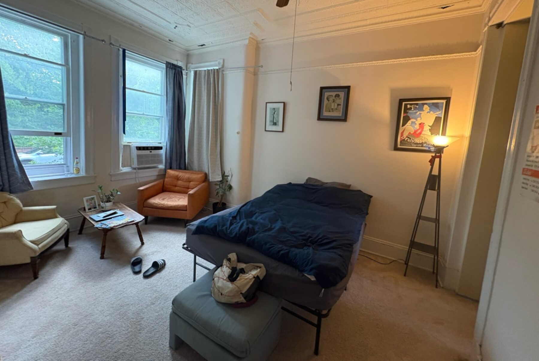 Brethren House Schlafzimmer: Bett mit dunkelblauer Decke, Stehlampe, zwei Sessel, Fenster mit Klimagerät.