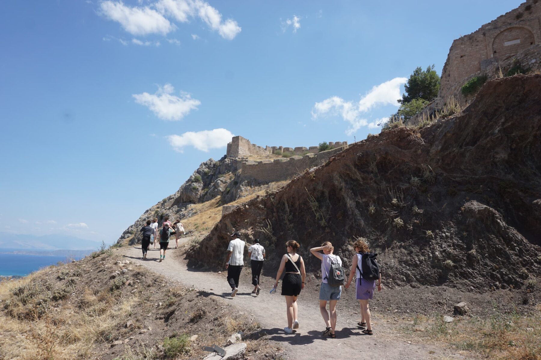 Wandergruppe auf Pfad zur Burgruine an steiler Küstenklippe in Griechenland, blaues Meer und klarer Himmel im Hintergrund