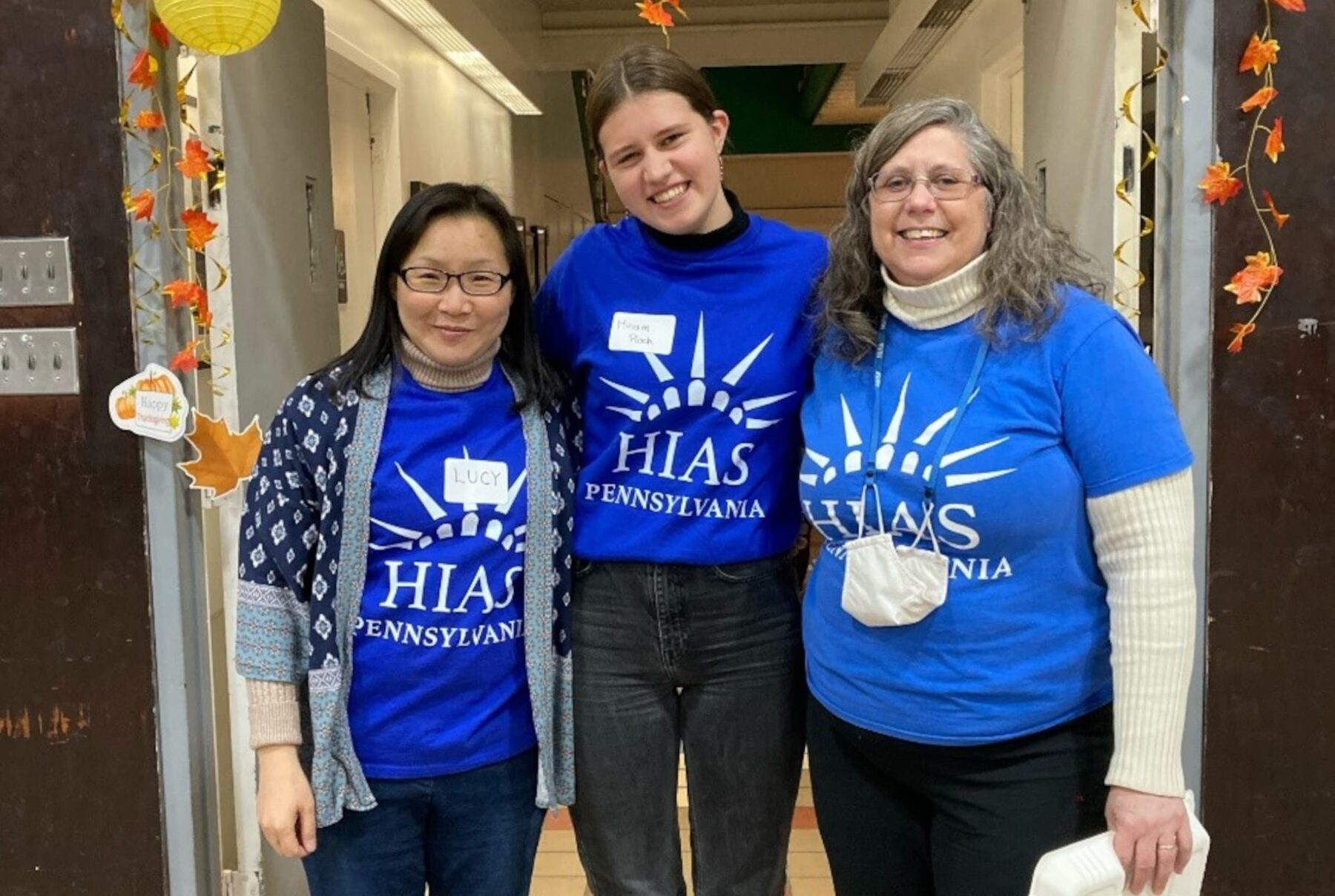 Drei lächelnde Freiwillige in blauen HIAS Pennsylvania-Shirts stehen eng beieinander im Flur mit Herbstdekoration.
