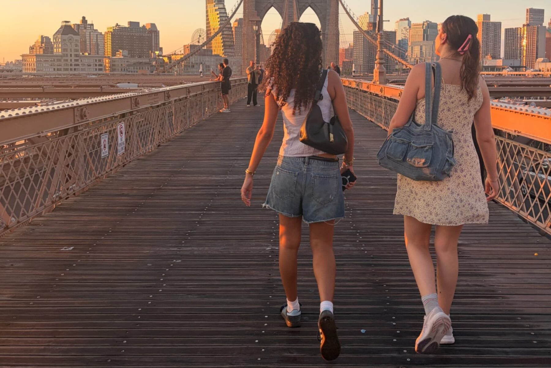 Zwei junge Frauen gehen auf der Brooklyn Bridge bei Sonnenuntergang, Holzplanken und Skyline im Hintergrund.