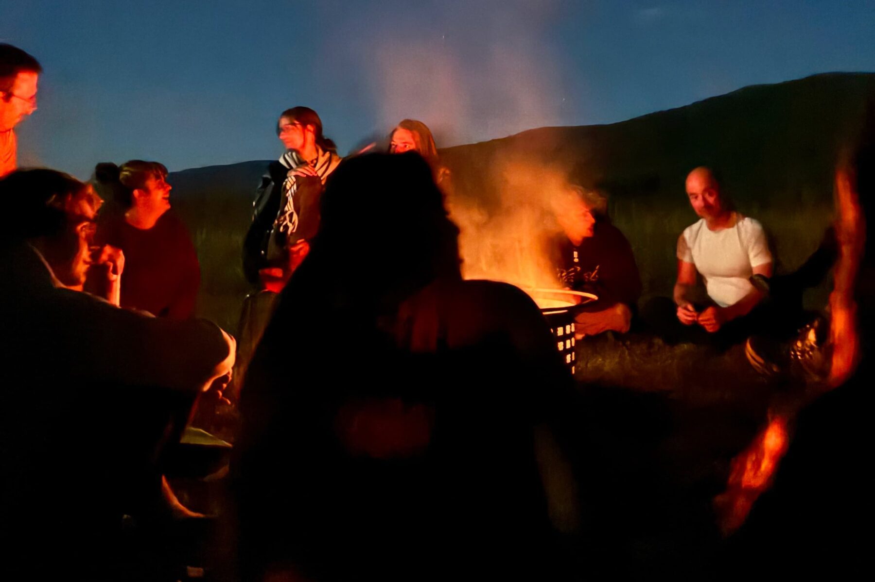Abendliches Lagerfeuer in Innisfree: Gruppe sitzt im Kreis um ein Feuer, orange Glut und Rauch, dunkle Silhouetten vor Hügeln.