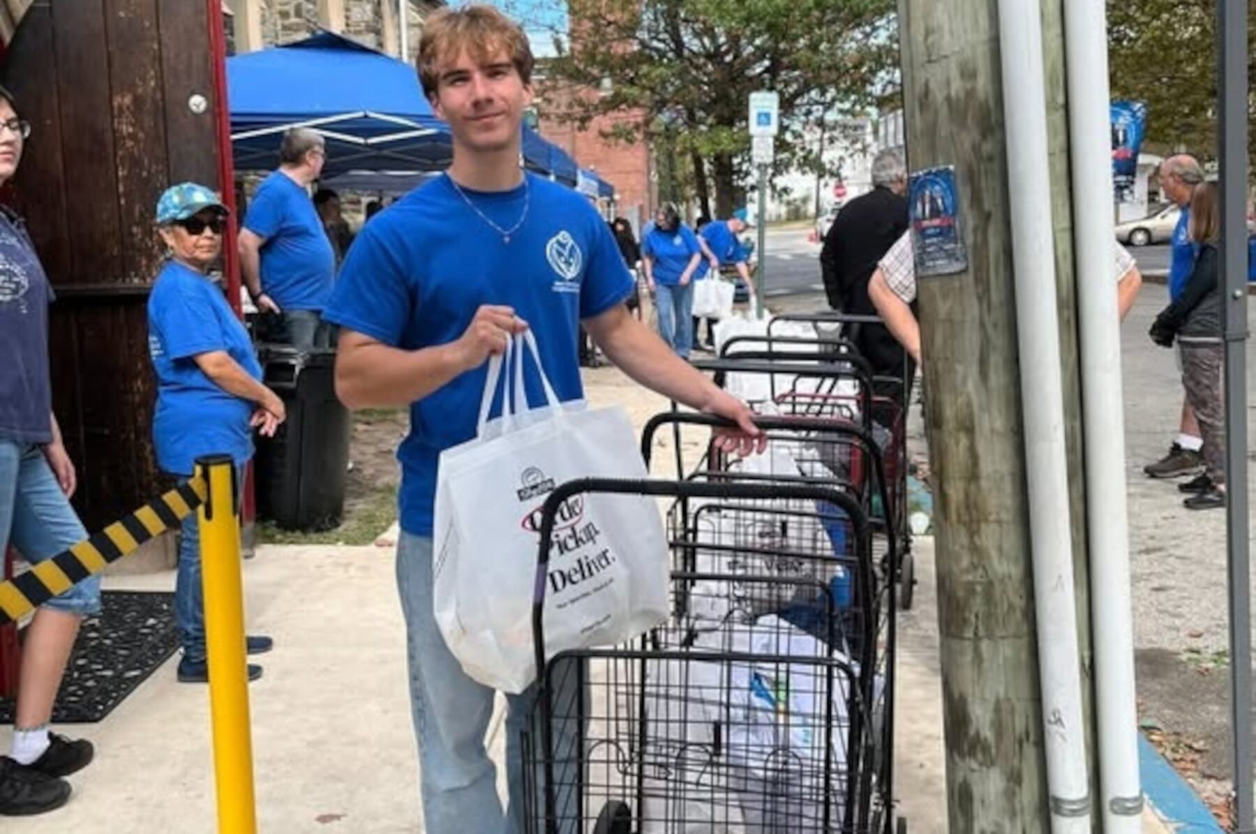 Junger Mann in blauem Shirt hält Tasche neben Einkaufswagen mit Spenden, Helfer im Hintergrund.