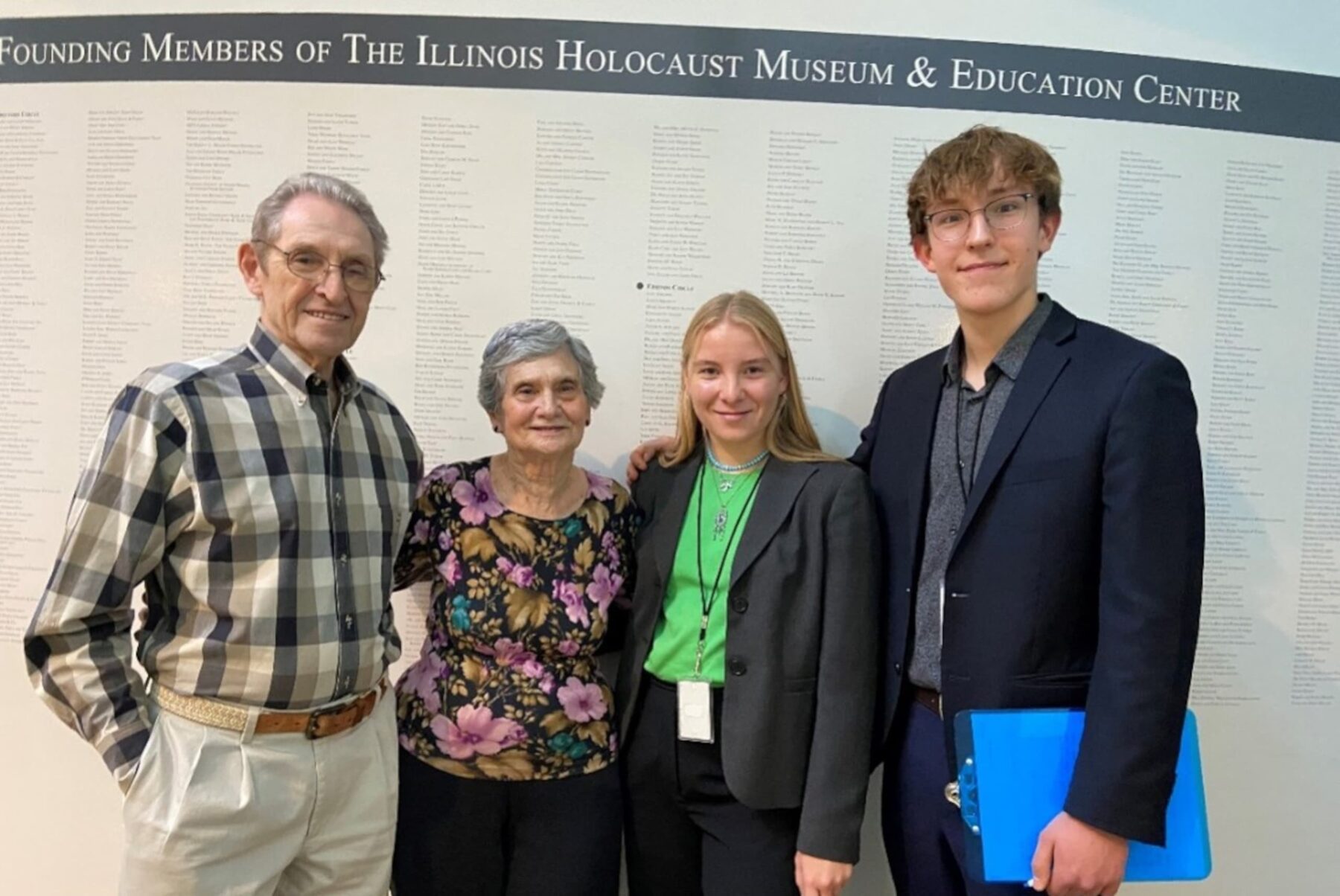 Vier Personen vor Wand mit Schriftzug des Illinois Holocaust Museum & Education Center, lächelnd, zwei Ältere und zwei junge Erwachsene.