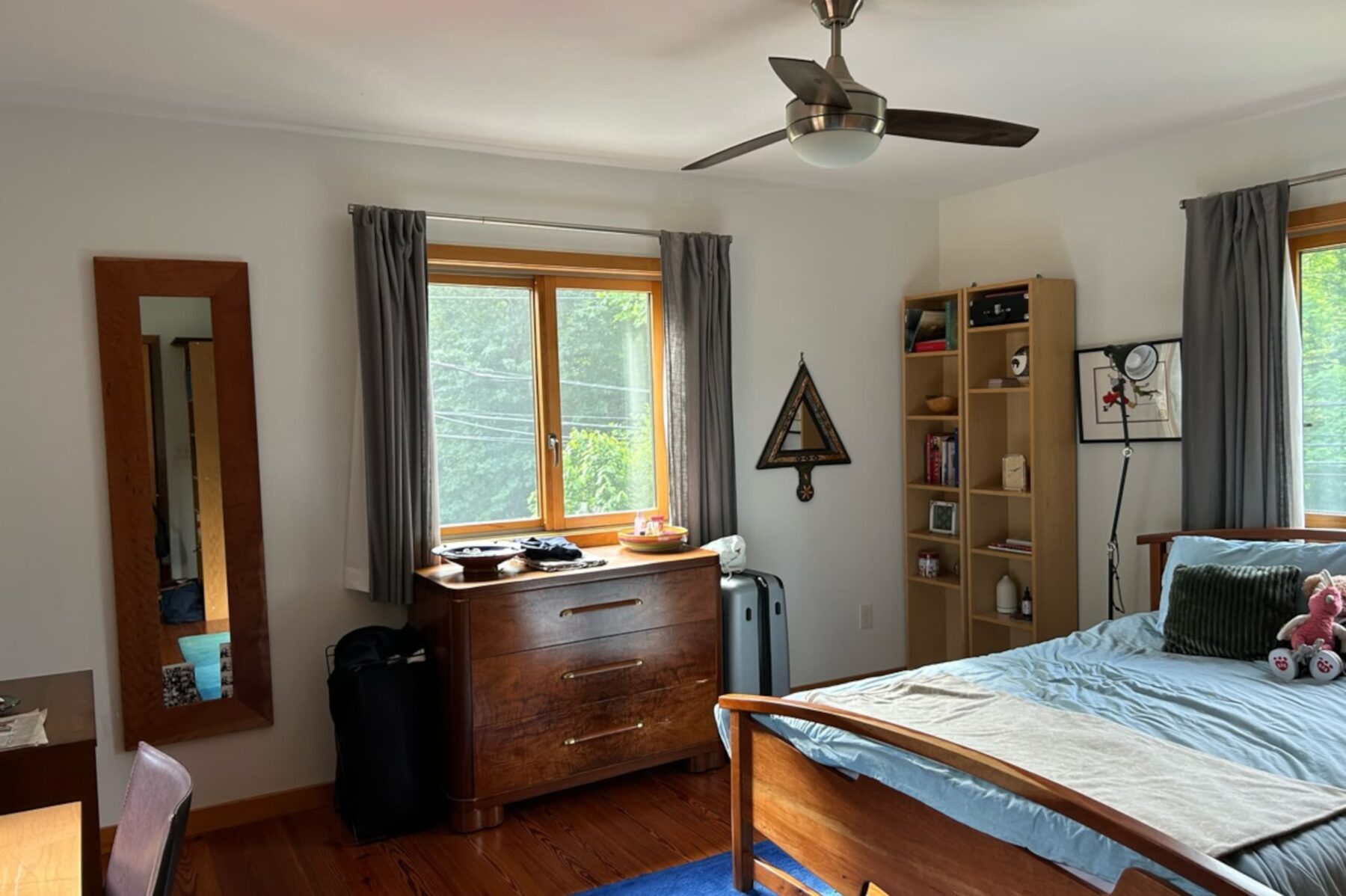 Helles Schlafzimmer mit Holzbett, blauem Bettzeug, Kommode unterm Fenster, Regal, Wandspiegel und Deckenventilator.