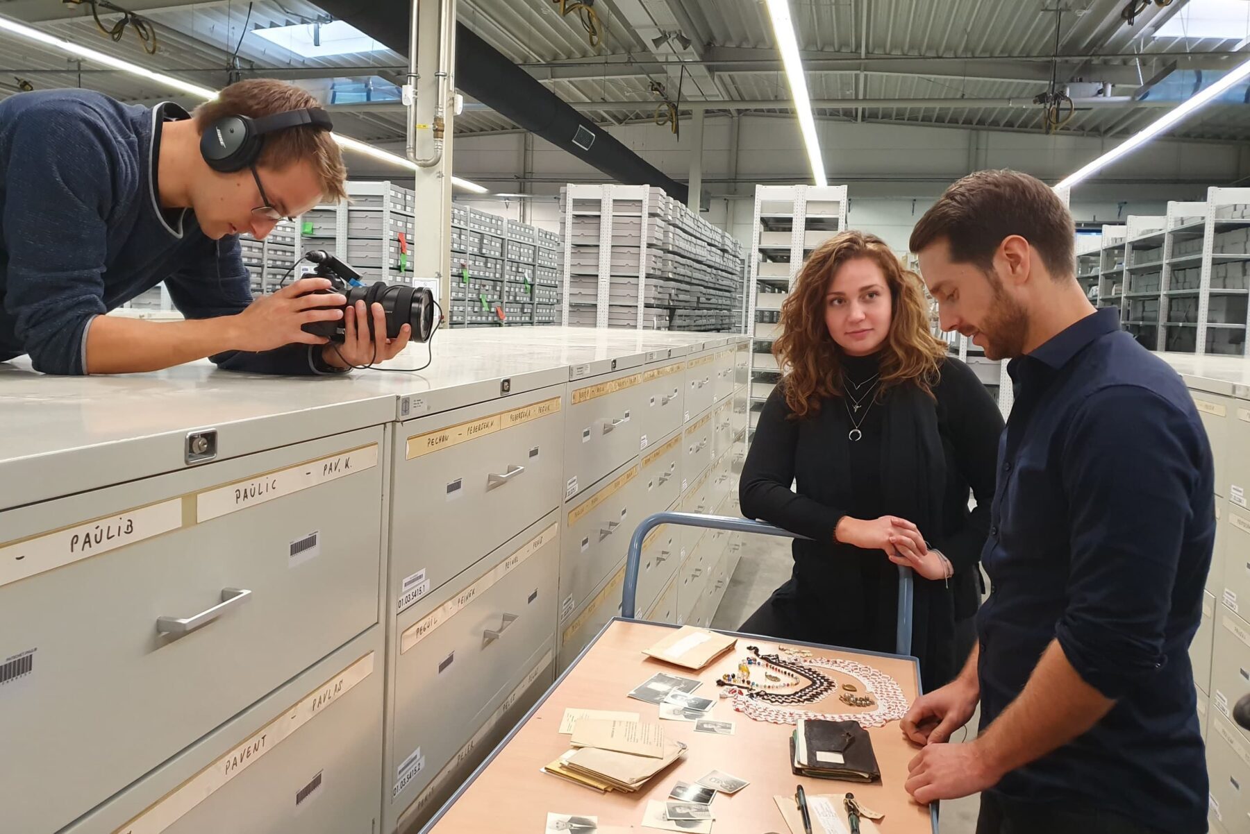Arolsen-Archiv: Kameramann filmt, zwei Personen begutachten Fotos, Briefe und Schmuck auf einem Rollwagen vor Archivschränken.