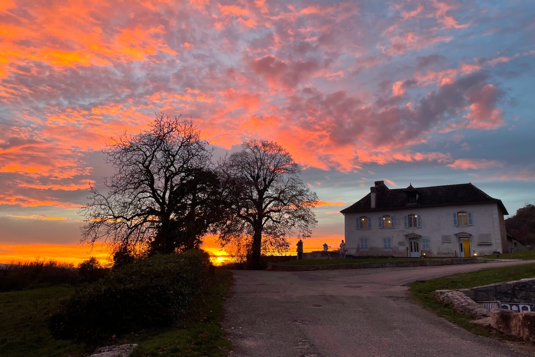 Maison in Izieu, Frankreich neben kahlen Bäumen, dramatischer orange-rosa Sonnenuntergangshimmel.
