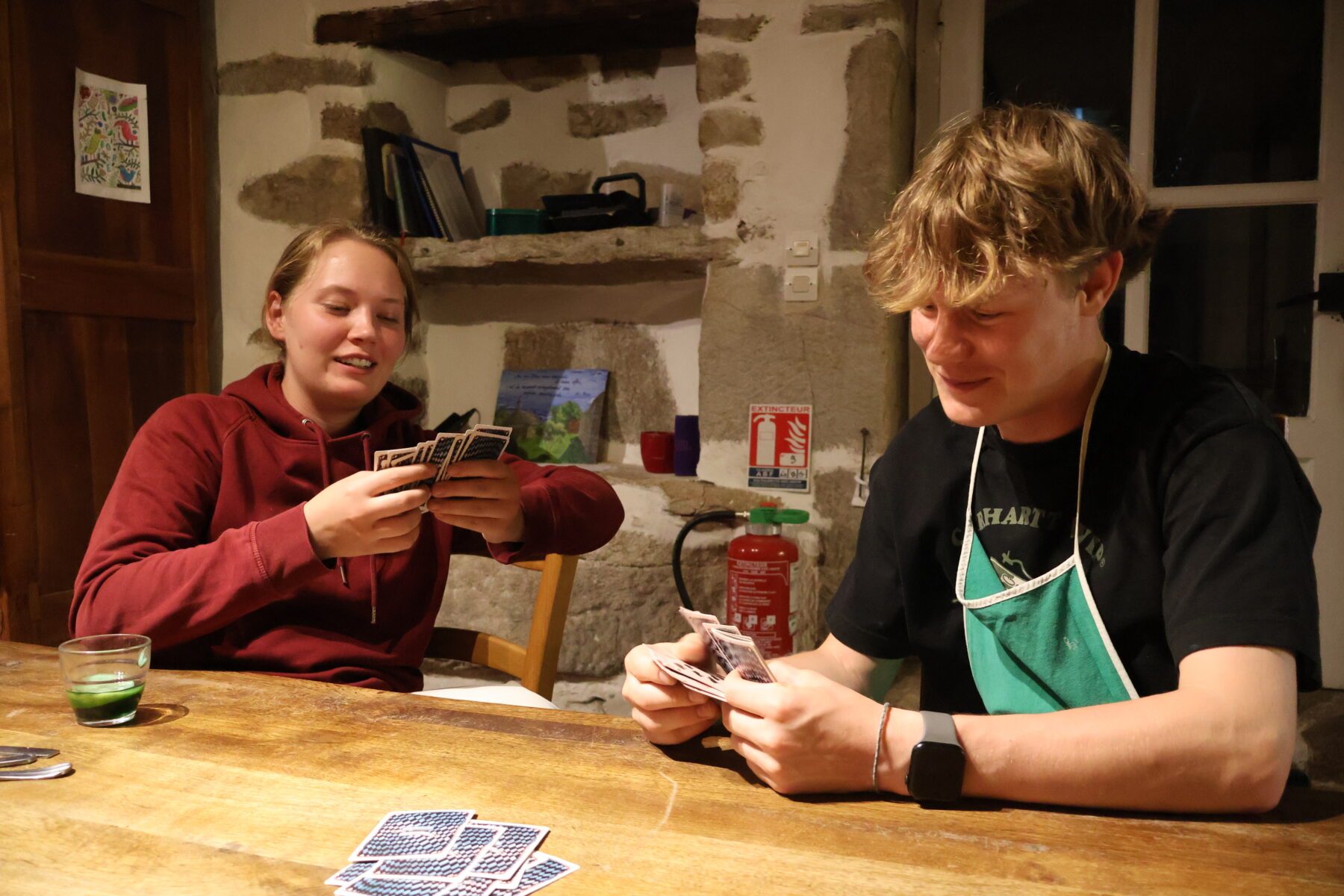 Zwei junge Leute spielen Karten an Holztisch in rustikaler Steinküche; Karten, Glas und Feuerlöscher sichtbar.
