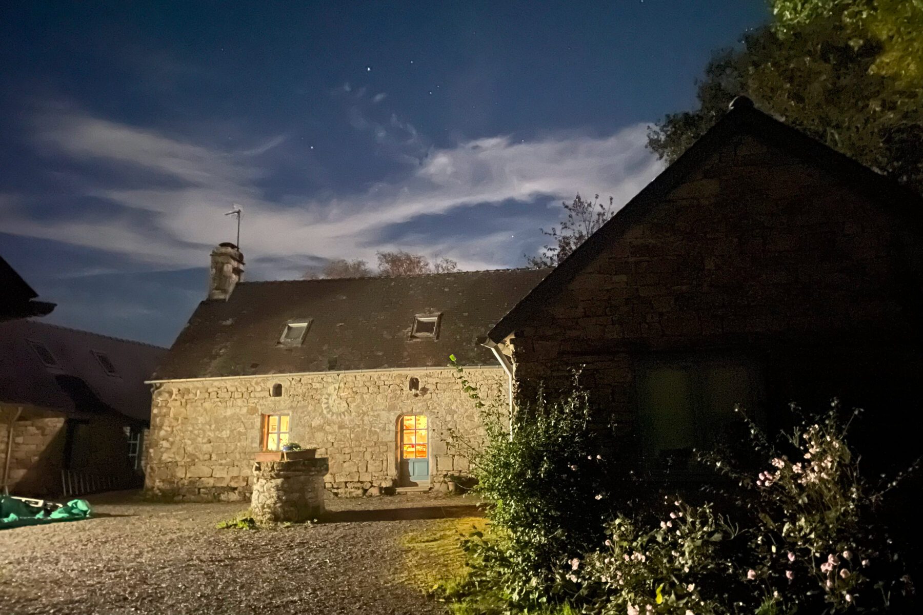 Steinhaus in Quimper bei Nacht, warm beleuchtete Tür und Fenster, Brunnen im Hof, Blumen rechts, wolkiger Sternenhimmel