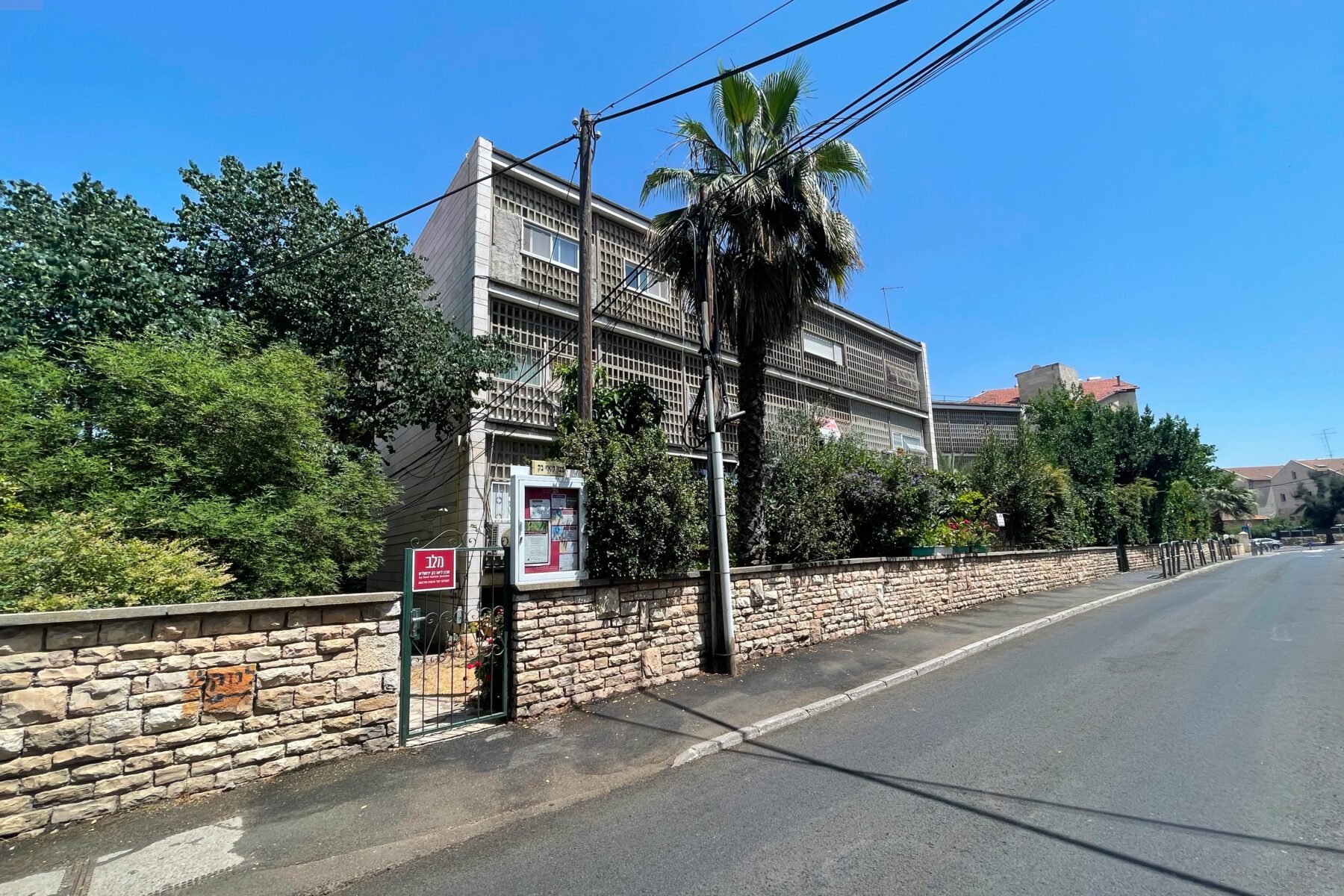 Leo-Baeck-Institut, Jerusalem: Außenansicht mit Rasterfassade, niedriger Sandsteinmauer, Palme, Tor und leerer Straße.