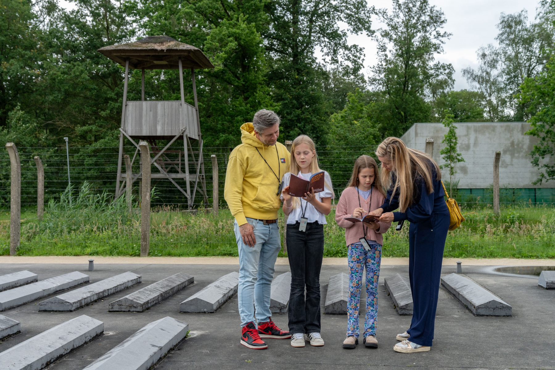 Vier Besucher am Gedenkort Kamp Vught lesen Informationsblätter zwischen Betonplatten vor einem hölzernen Wachturm