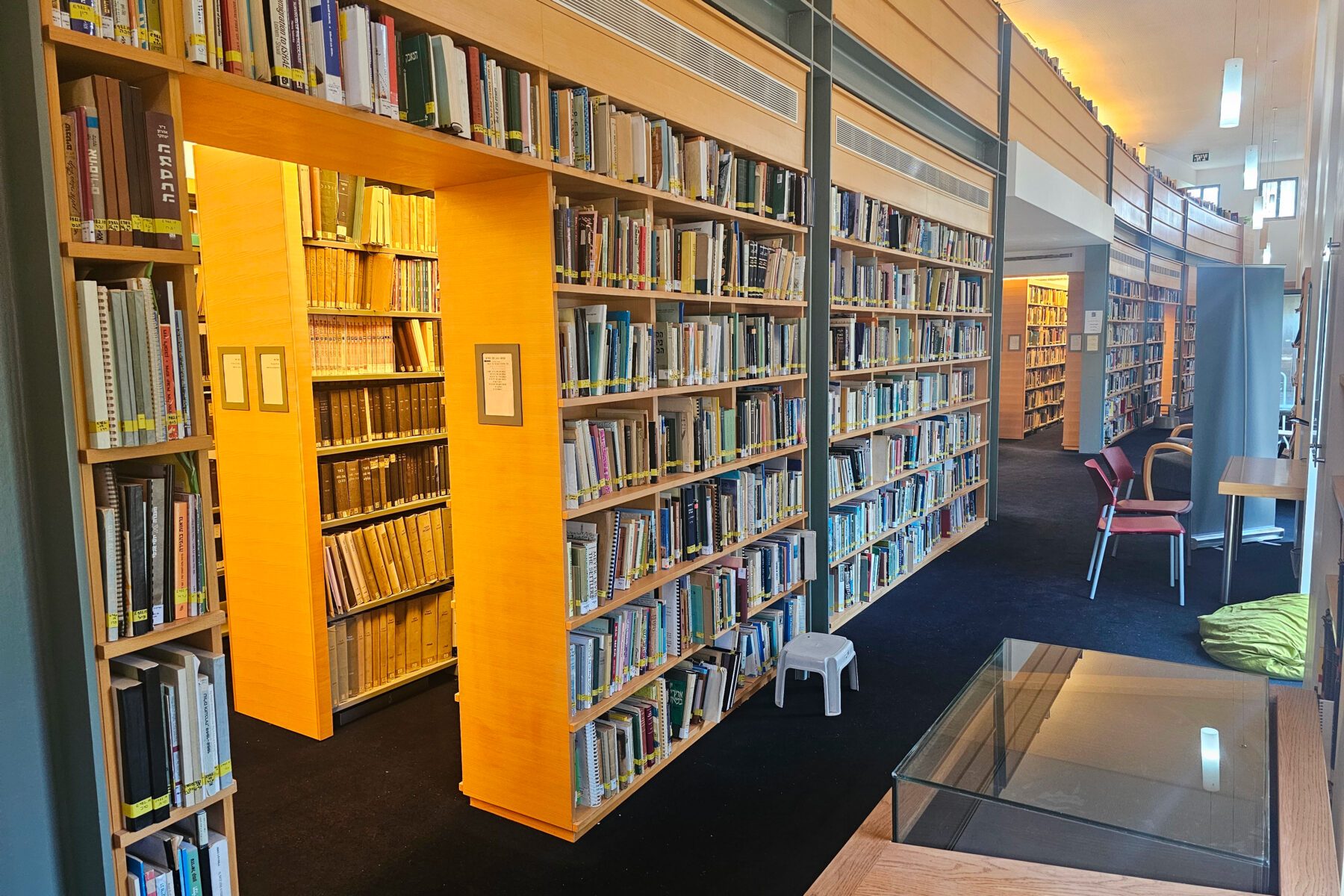 Innenraum der Bibliothek Yad Ben Zvi in Jerusalem: lange helle Holzregale mit Büchern, Gang, Stuhl, Hocker und Glasvitrine.
