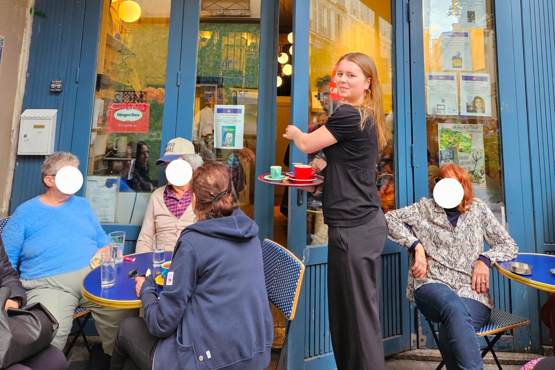 Kellnerin trägt rote und grüne Tassen zu Gästen an Außentischen vor blauem Café des Psaumes, Frankreich.
