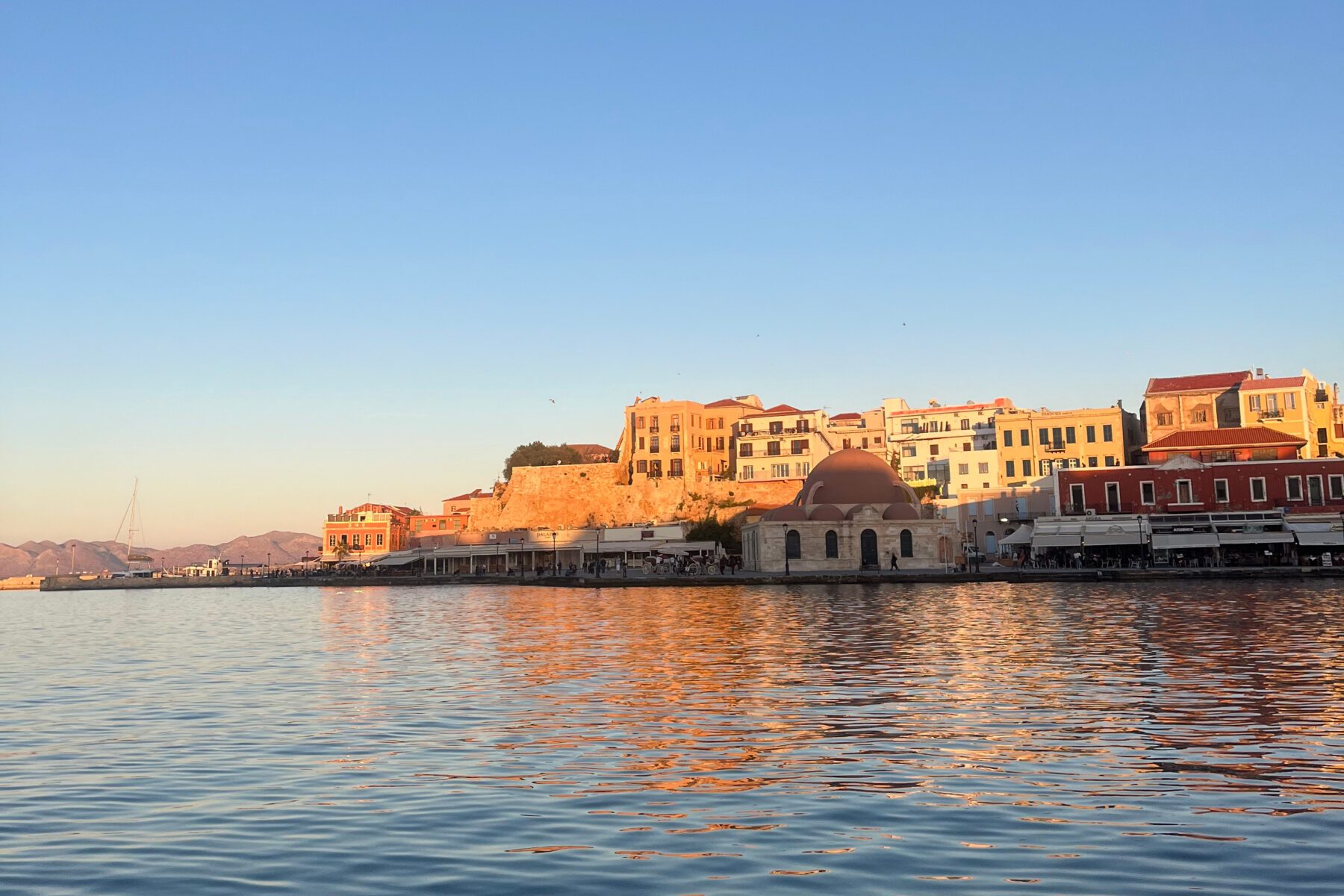 Chania/Hafen in Griechenland bei Sonnenuntergang: goldene Hafenbauten und Kuppelmoschee spiegeln sich im ruhigen Wasser.