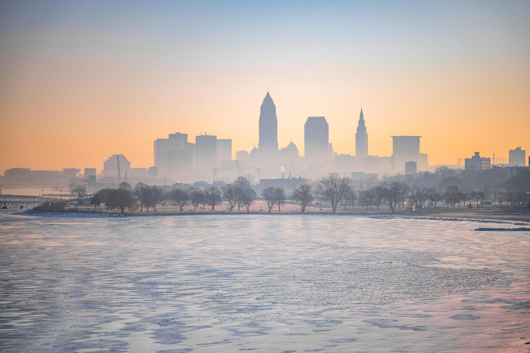 Cleveland Skyline im Dunst bei Sonnenaufgang über dem zugefrorenen Lake Erie, Baumreihe am Ufer sichtbar.