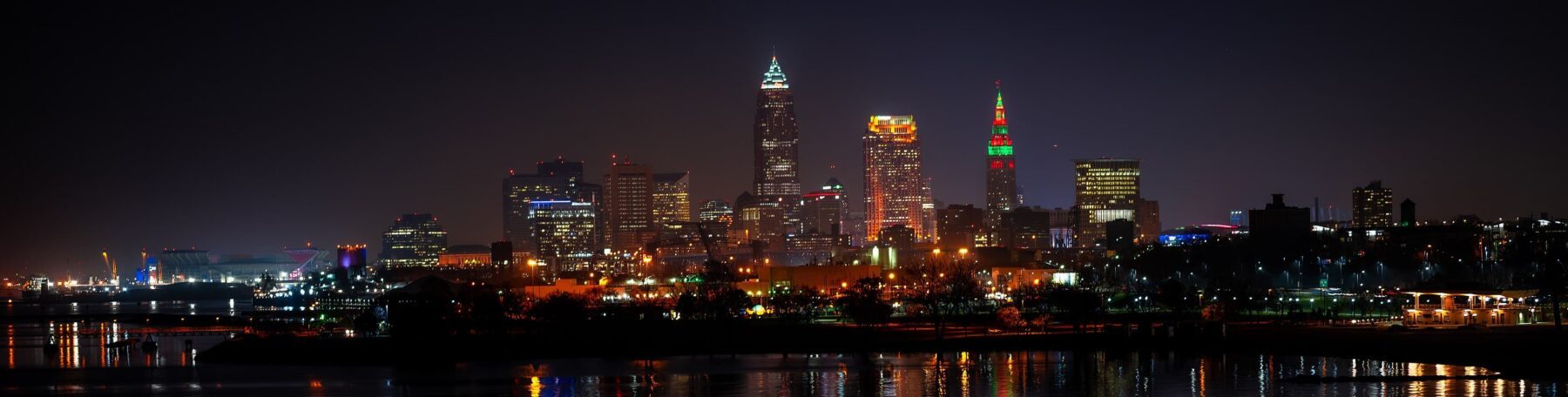 Cleveland-Skyline bei Nacht, leuchtende Wolkenkratzer inkl. bunt beleuchtetem Terminal Tower und Lichterspiegelung im Wasser