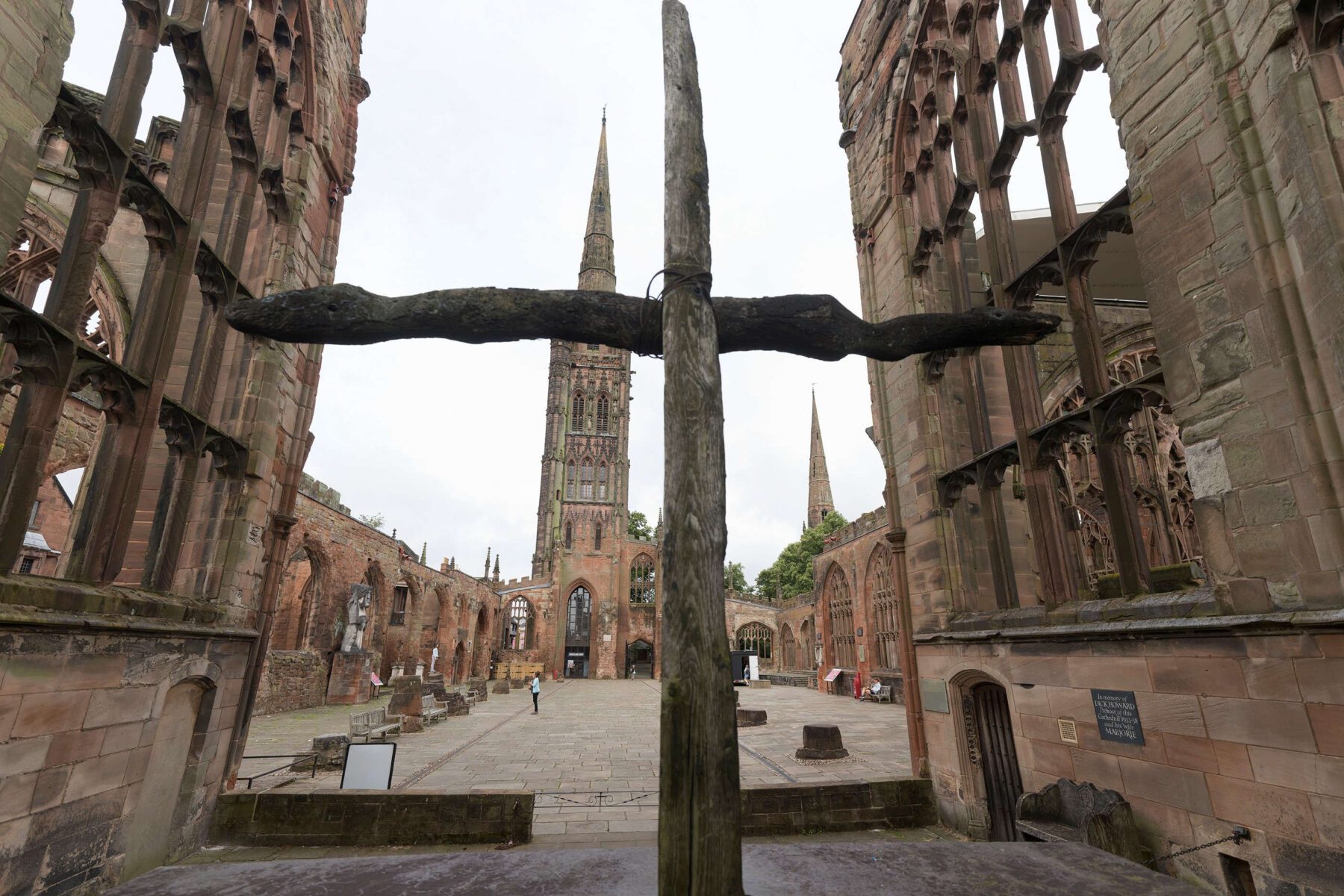 Holzkreuz im Vordergrund vor den verfallenen Mauern und Türmen der Coventry Cathedral, offener Innenhof, wolkiger Himmel
