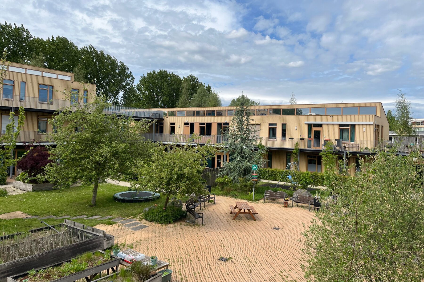 Innenhof Dagcentrum Maccabiadelaan, Amsterdam: Holzbau, gepflasterter Platz mit Tisch, Bänken, Bäumen und Beeten.