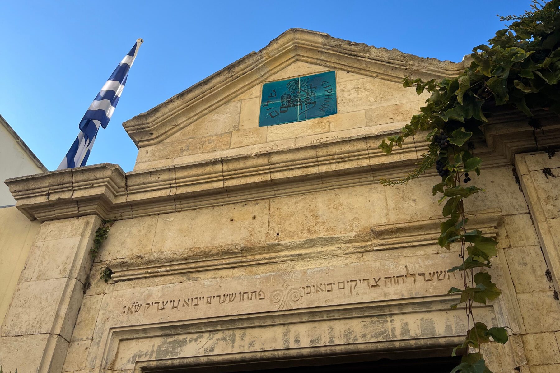 Etz Hayyim-Synagoge in Chania: Steinfassade mit hebräischer Inschrift, blauem Emblem, griechischer Fahne und Weinranke.