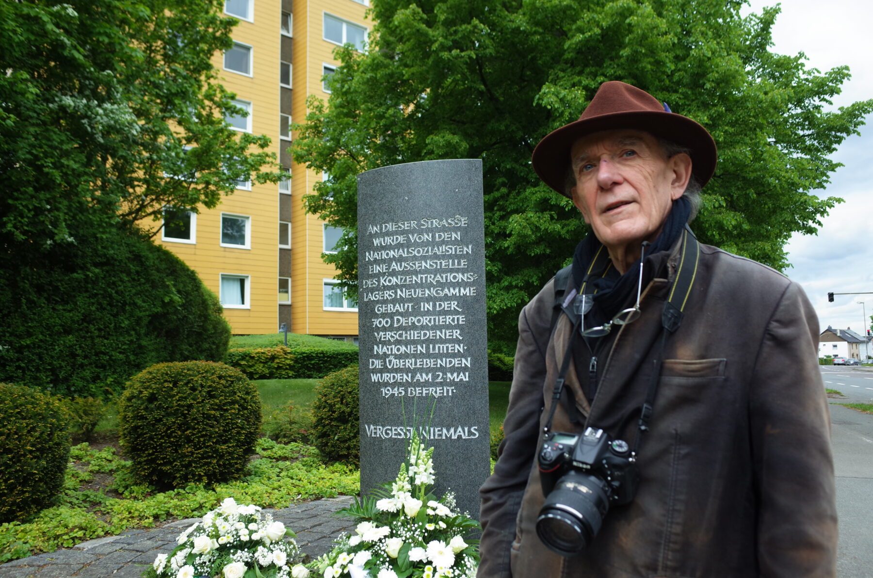 Älterer Mann mit Filzhut und Kamera vor Gedenkstein mit Blumenkränzen, Wohnhaus und Bäumen im Hintergrund.