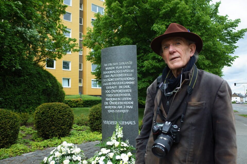 Älterer Mann mit Filzhut und Kamera vor Gedenkstein mit Blumenkränzen, Wohnhaus und Bäumen im Hintergrund.