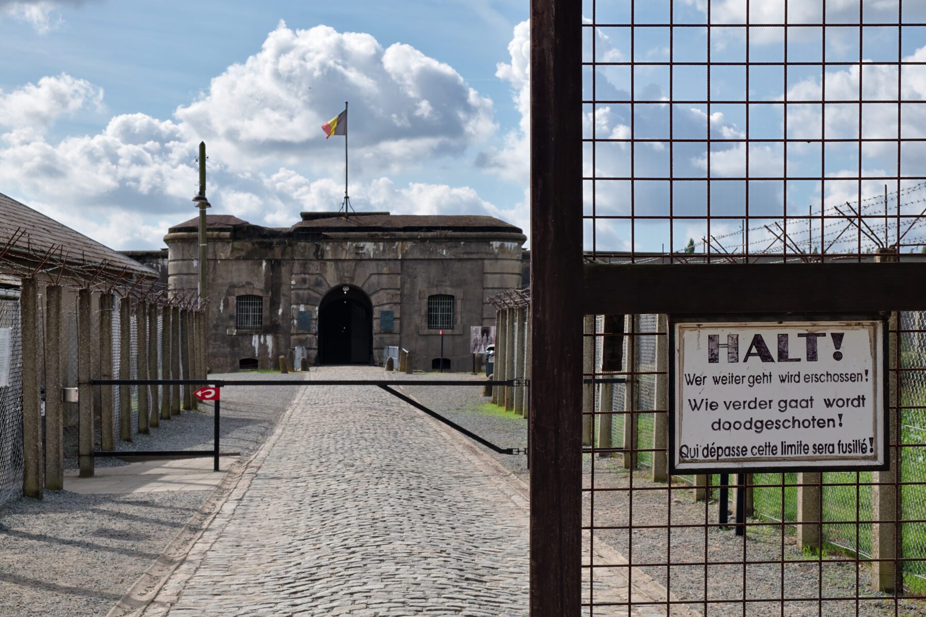 Eingang Fort Breendonk: gepflasterter Weg zur Toranlage mit Stacheldraht, Warnschild 'HALT!' und belgischer Flagge