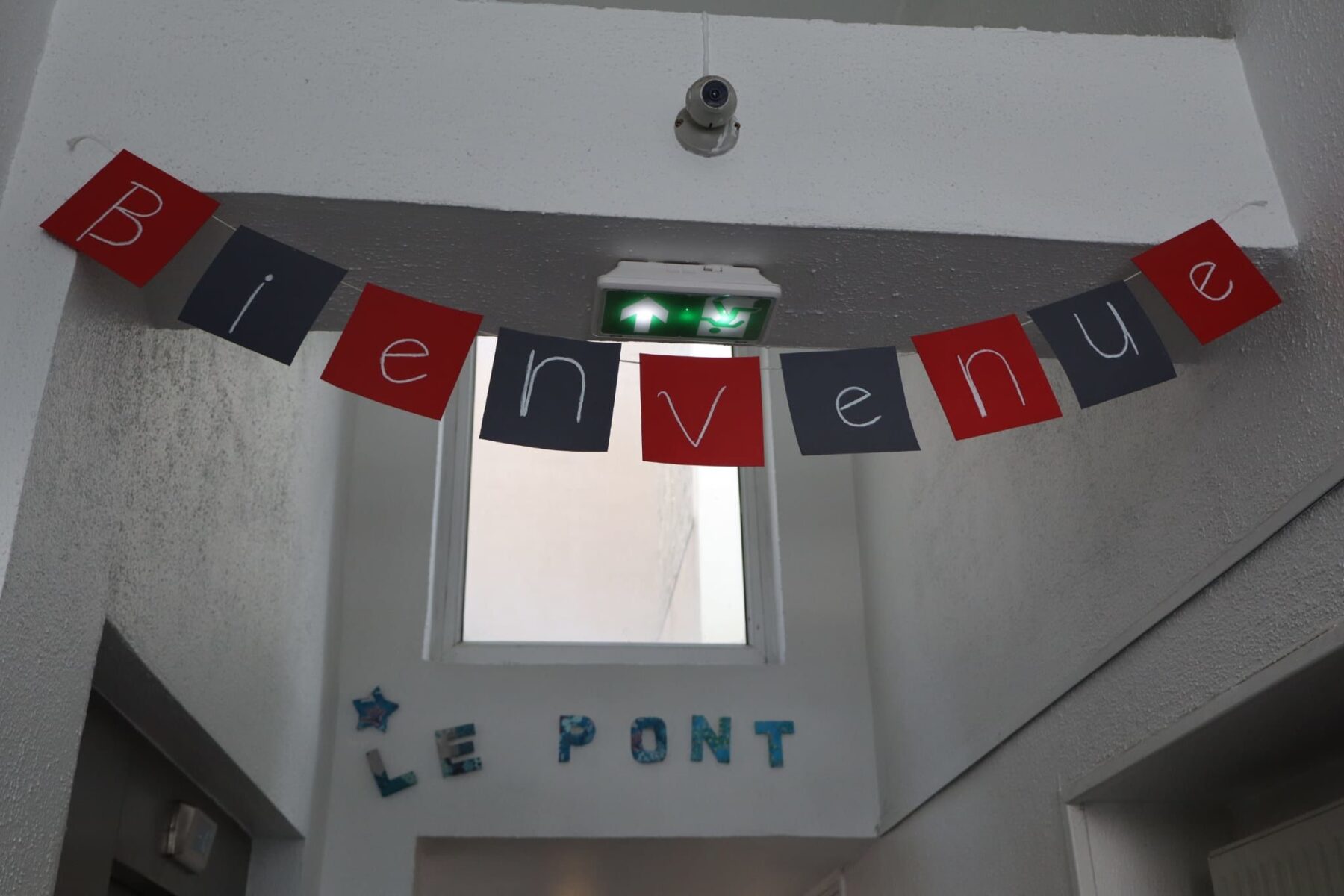 Foyer 'Le Pont' mit hängender Girlande 'Bienvenue' über Fenster und grünem Notausgangsschild.