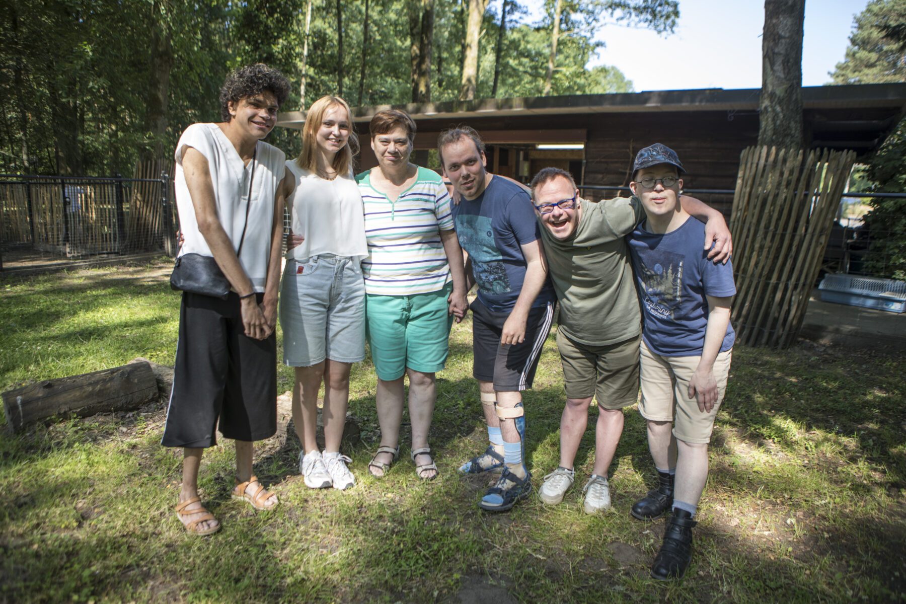 Sieben Freiwillige der Arche Monnikenheide stehen Arm in Arm vor einer Holzhütte im Wald.
