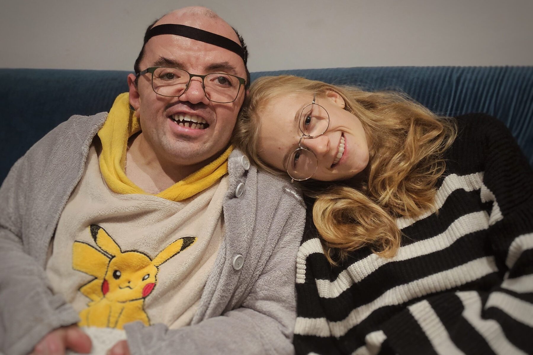 Zwei ASF‑Freiwillige auf Sofa, eine im Pikachu‑Poncho und grauem Bademantel, die andere im schwarz‑weißen Streifenpullover.