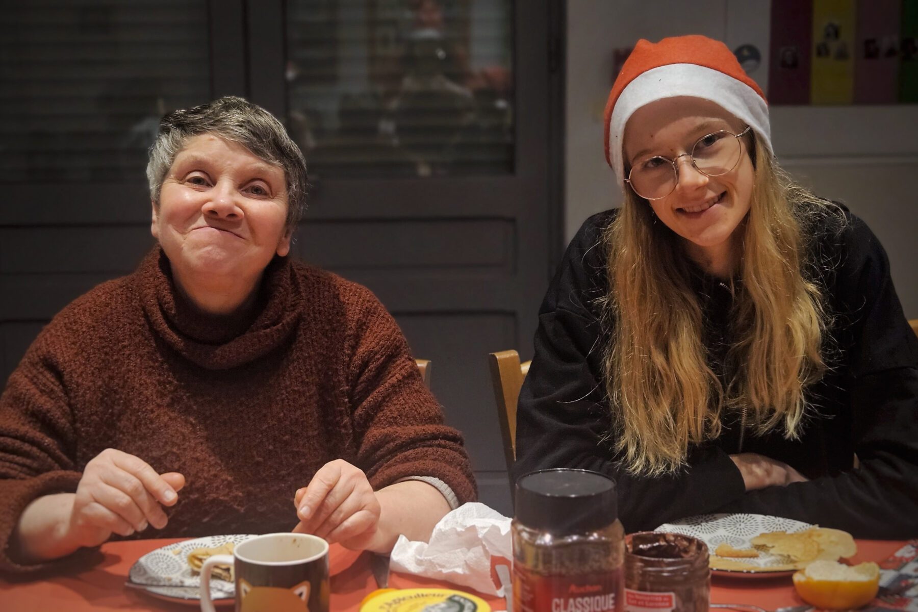 Zwei Freiwillige der Arche in Lille an Tisch; links brauner Pullover, rechts Nikolausmütze, Tasse, Aufstrich.
