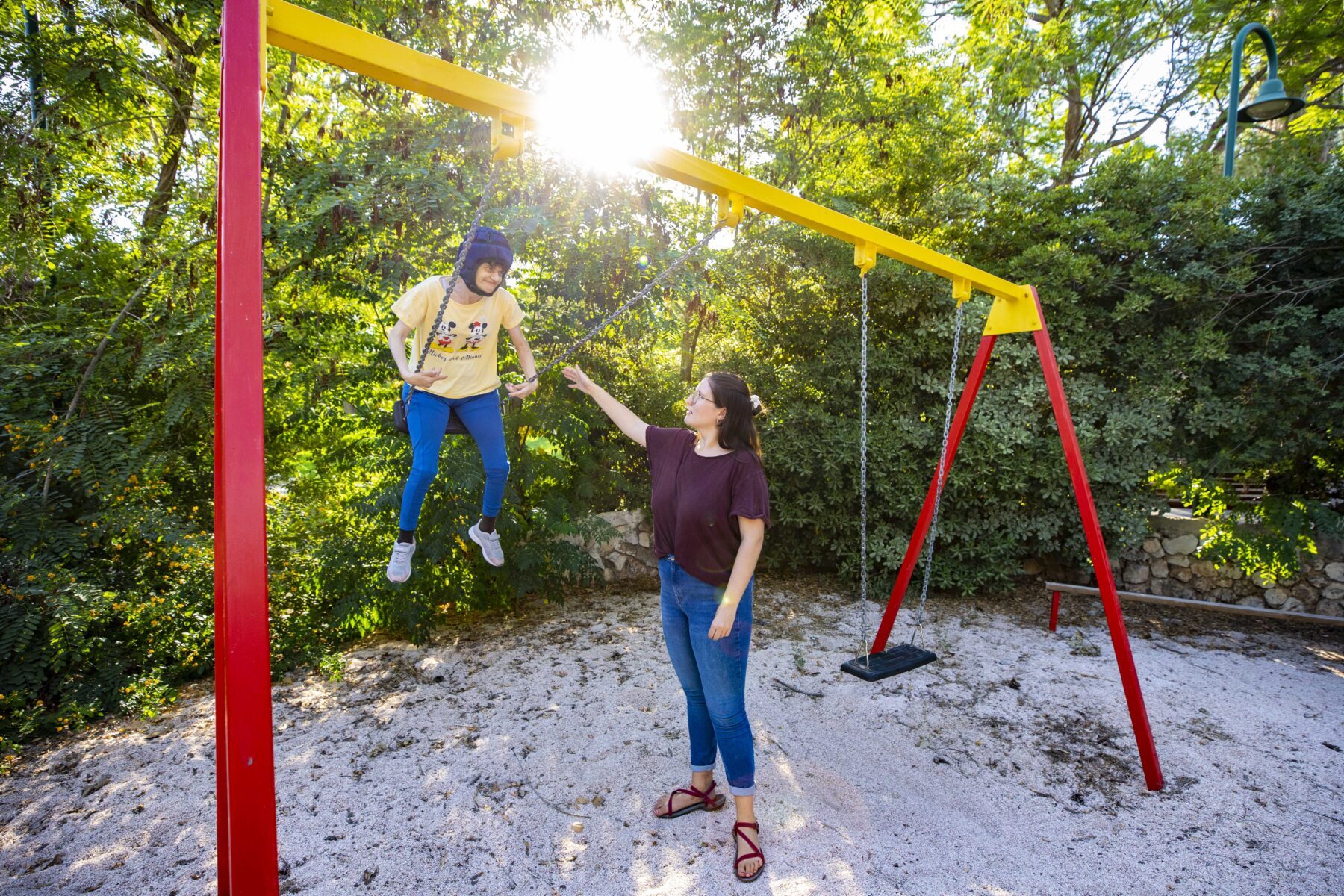 ASF-Freiwillige schiebt Kind mit Helm auf roter-gelber Schaukel im sonnigen Park vor Bäumen.