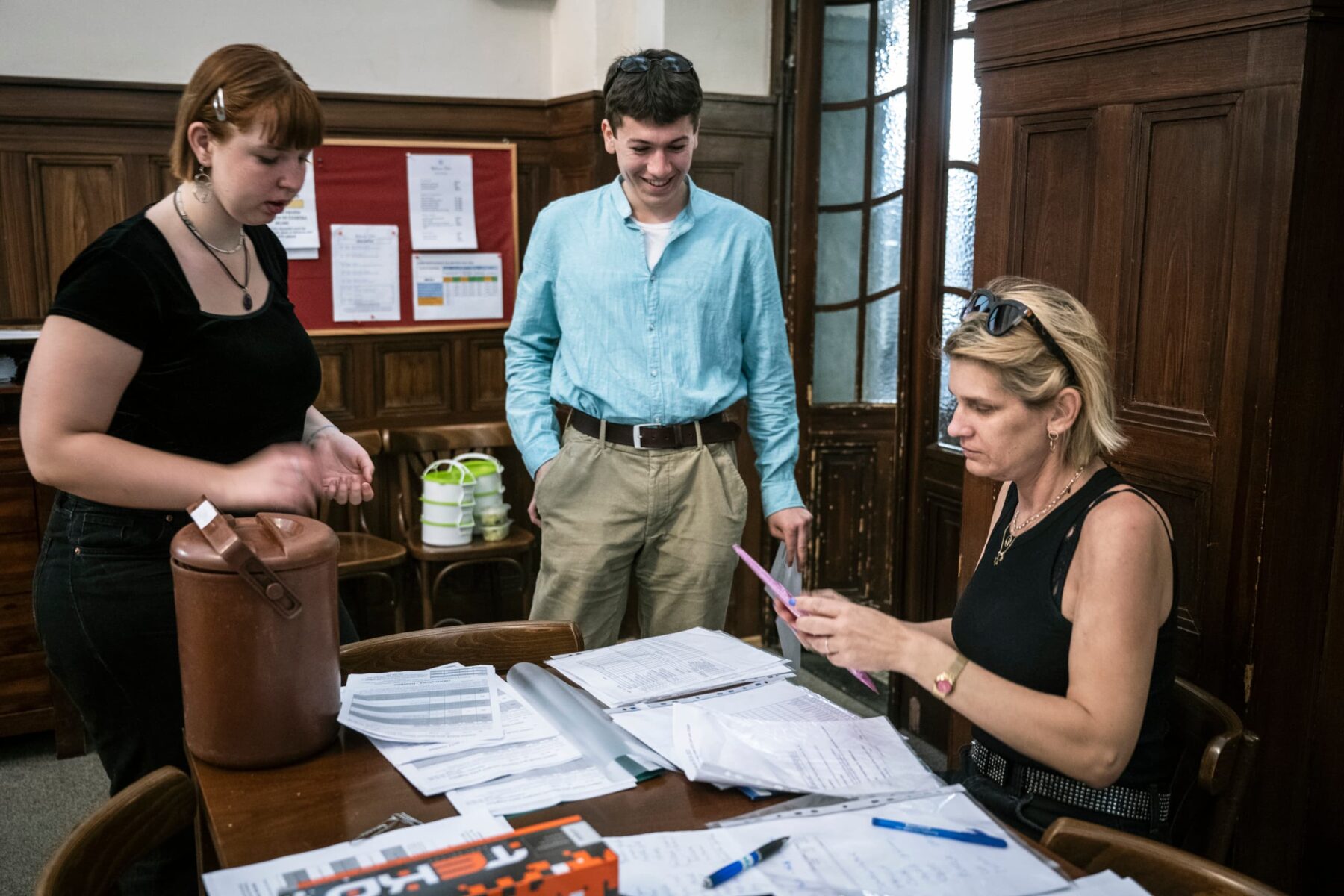 Drei Freiwillige in Prag an einem Tisch mit Formularen; eine Frau sortiert Dokumente, zwei junge Leute stehen daneben.