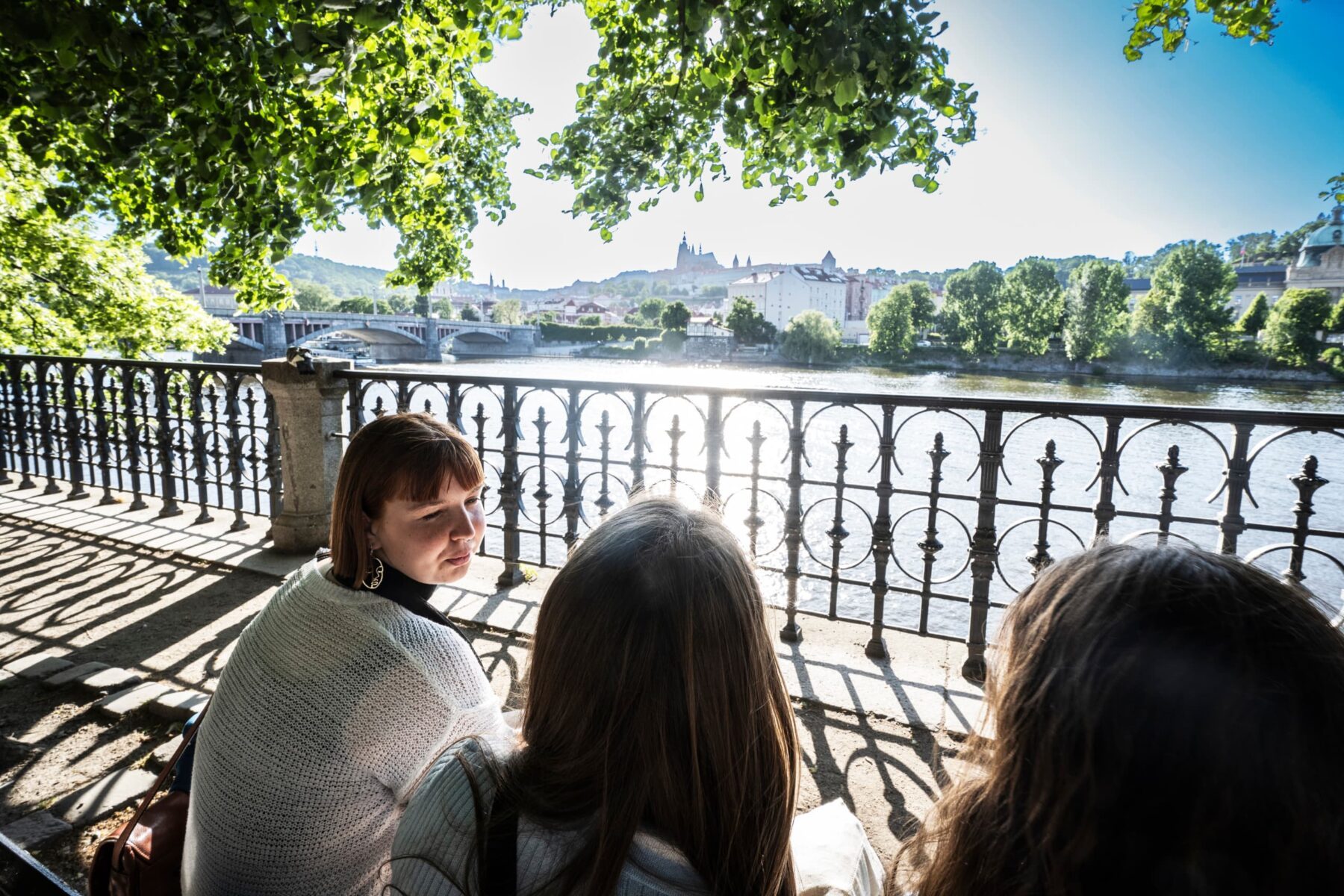 Drei ASF‑Freiwillige sitzen am Moldauufer unter Bäumen, schmiedeeisernes Geländer, Sonne spiegelt, Prager Burg im Hintergrund