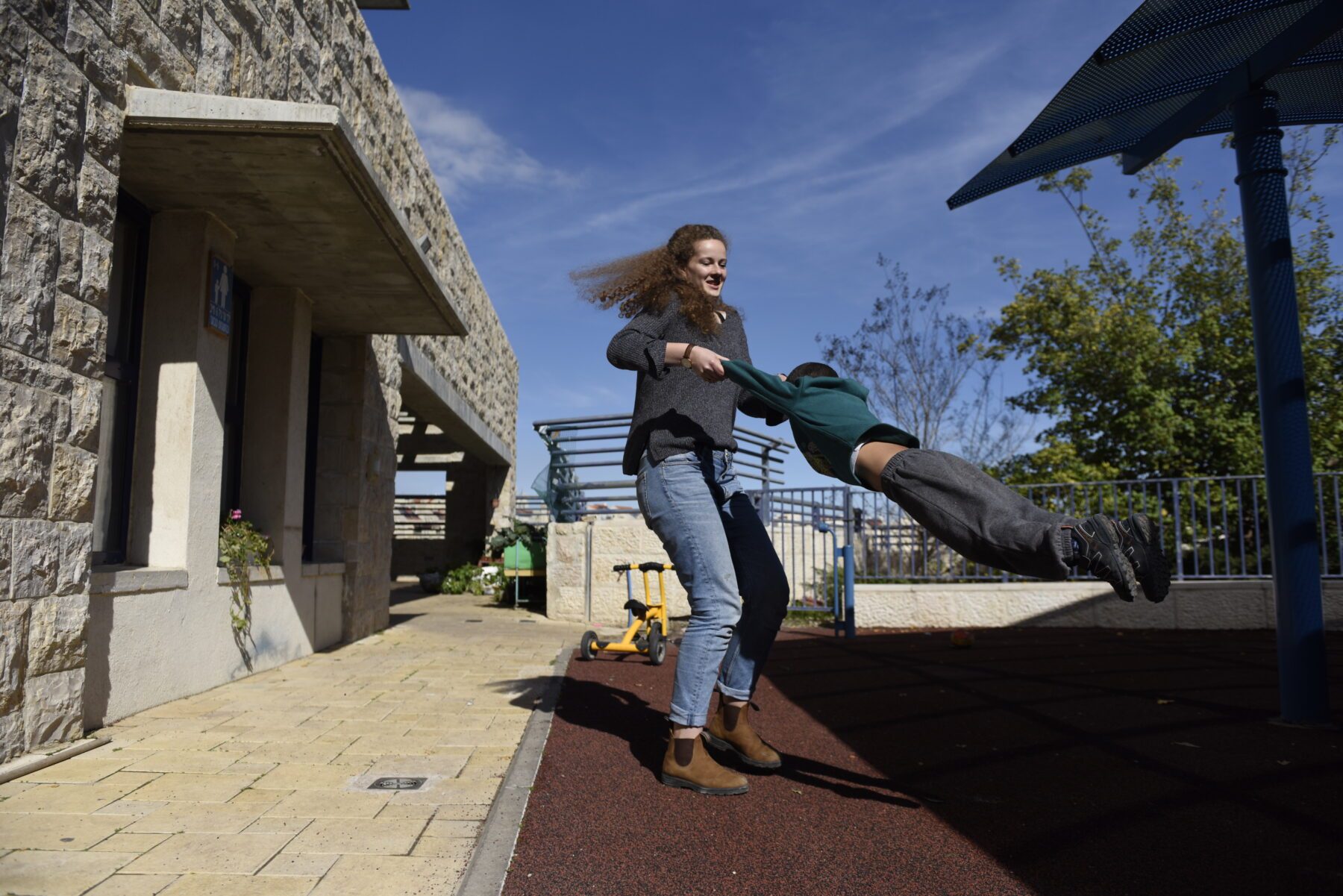 Freiwillige schwingt ein Kind an den Armen auf Spielplatz in Jerusalem; Steinmauer, blauer Himmel.
