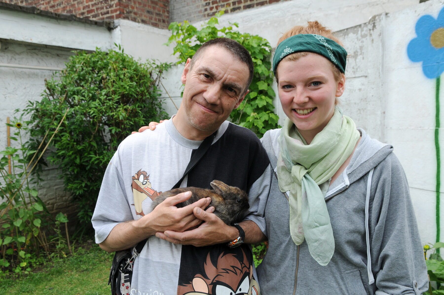 Mann hält braunes Kaninchen im Arm, junge Frau mit grünem Bandana lächelnd neben ihm, Gartenwand mit Blumenmalerei.