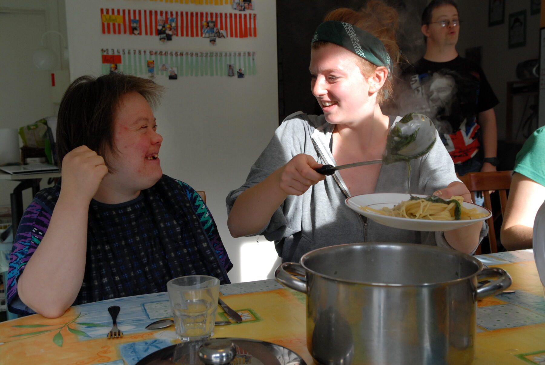 Freiwillige schöpft dampfende Spaghetti mit Kelle aus großem Topf und serviert sie Kindern an einem bunt gedeckten Esstisch.