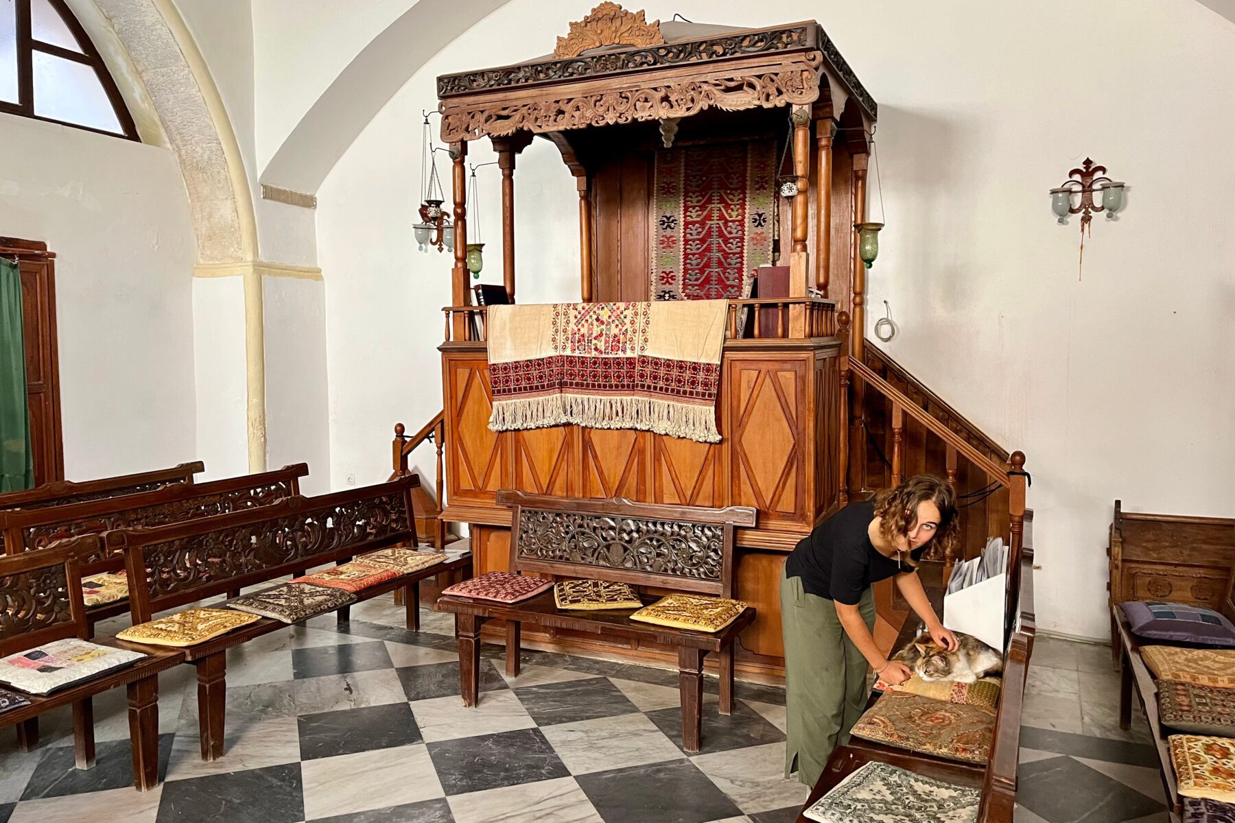 Etz Hayyim Synagoge (Chania): geschnitztes Holzpult mit Teppich, schwarz-weißes Marmormosaik, Freiwillige streichelt Katze.
