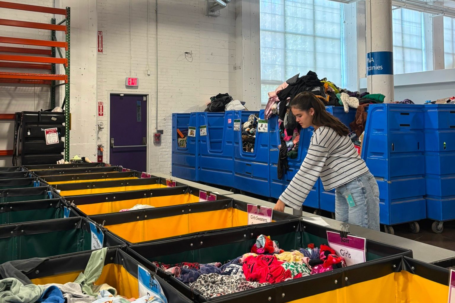 Freiwillige sortiert Kinderkleidung in gelben und grünen Wannen vor blauen Rollcontainern in Lagerhalle.