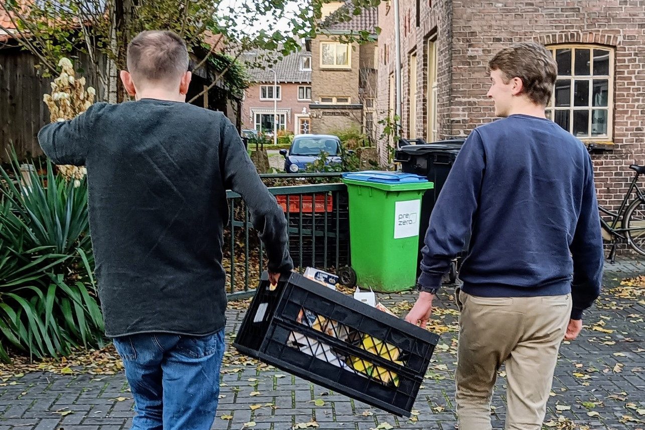 Zwei Freiwillige tragen eine schwarze Kiste mit Müllverpackungen durch eine Pflastergasse vor Häusern in Gouda.