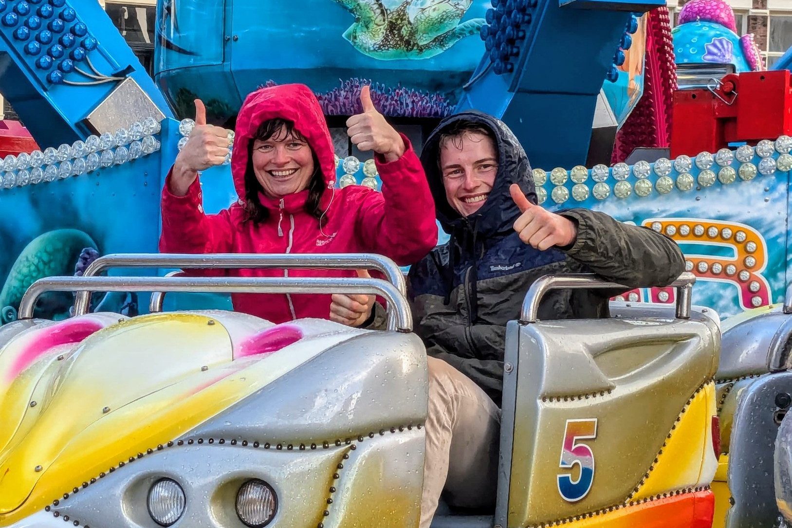 Zwei Freiwillige in Regenjacken auf buntem Kirmeswagen Nr.5 in Gouda, Kapuzen hoch, Daumen hoch, Wassertropfen auf Lack
