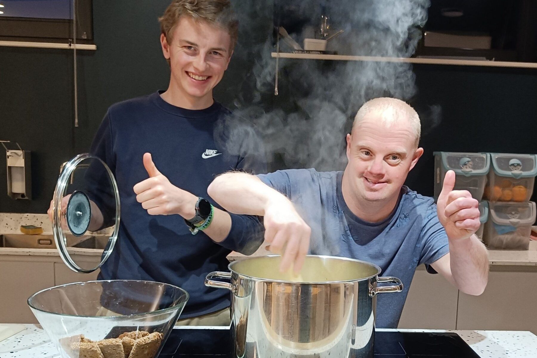 Zwei Freiwillige in der Ark Gouda (Niederlande) kochen an dampfendem Topf, einer hält Deckel, beide zeigen Daumen hoch.