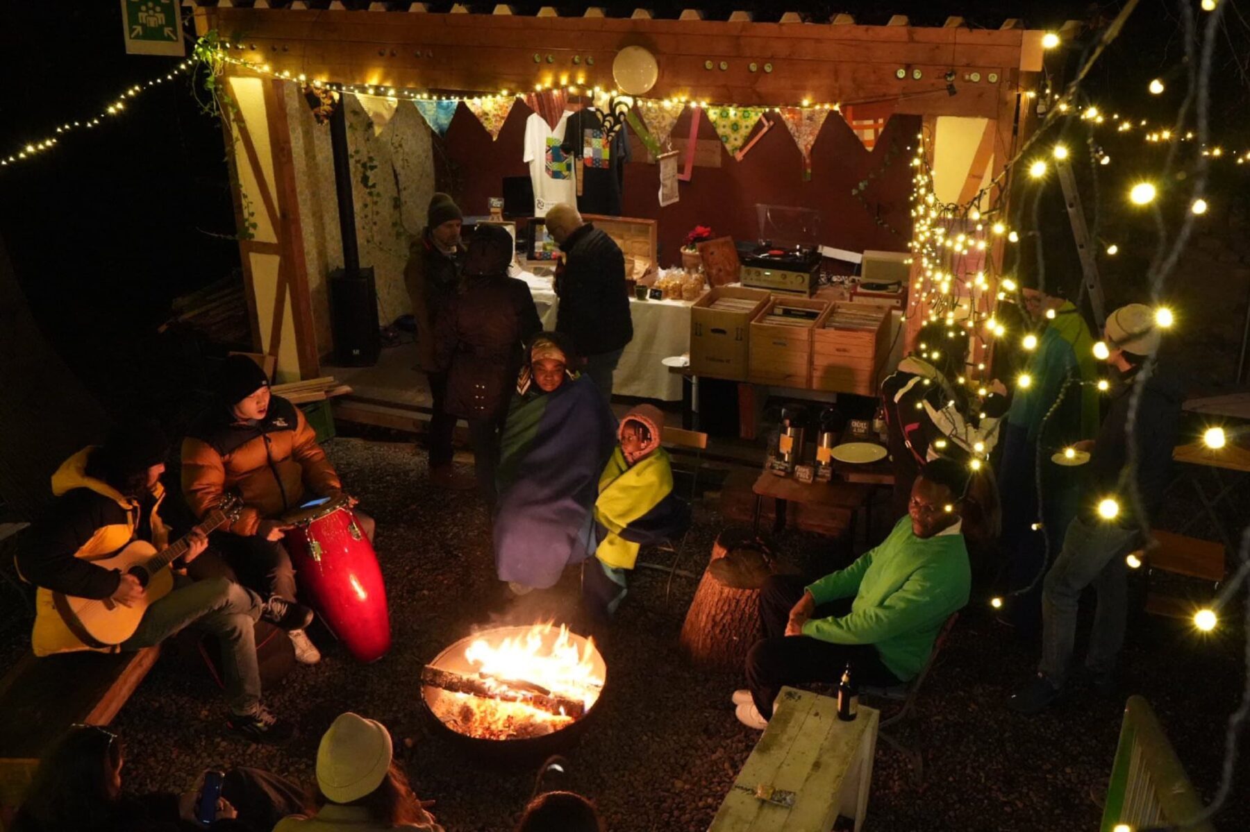 Freiwillige am Grandhotel-Stand beim Feuermarkt Augsburg sitzen um ein Feuer, Lichterketten, Musiker mit Gitarre und Trommel.