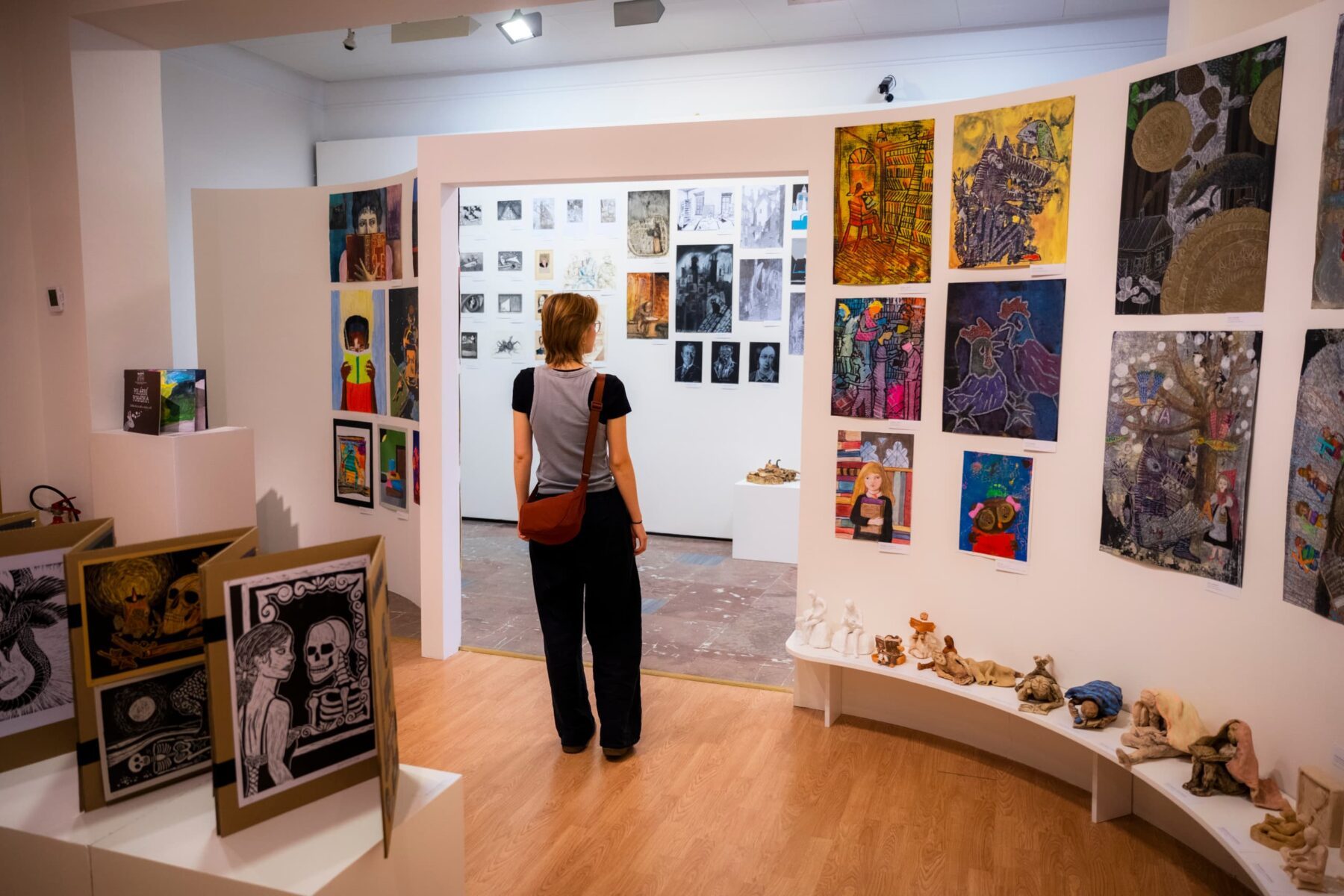Person mit brauner Umhängetasche betrachtet bunte Bilder und Tonfiguren in einer Ausstellung in der Gedenkstätte Lidice.