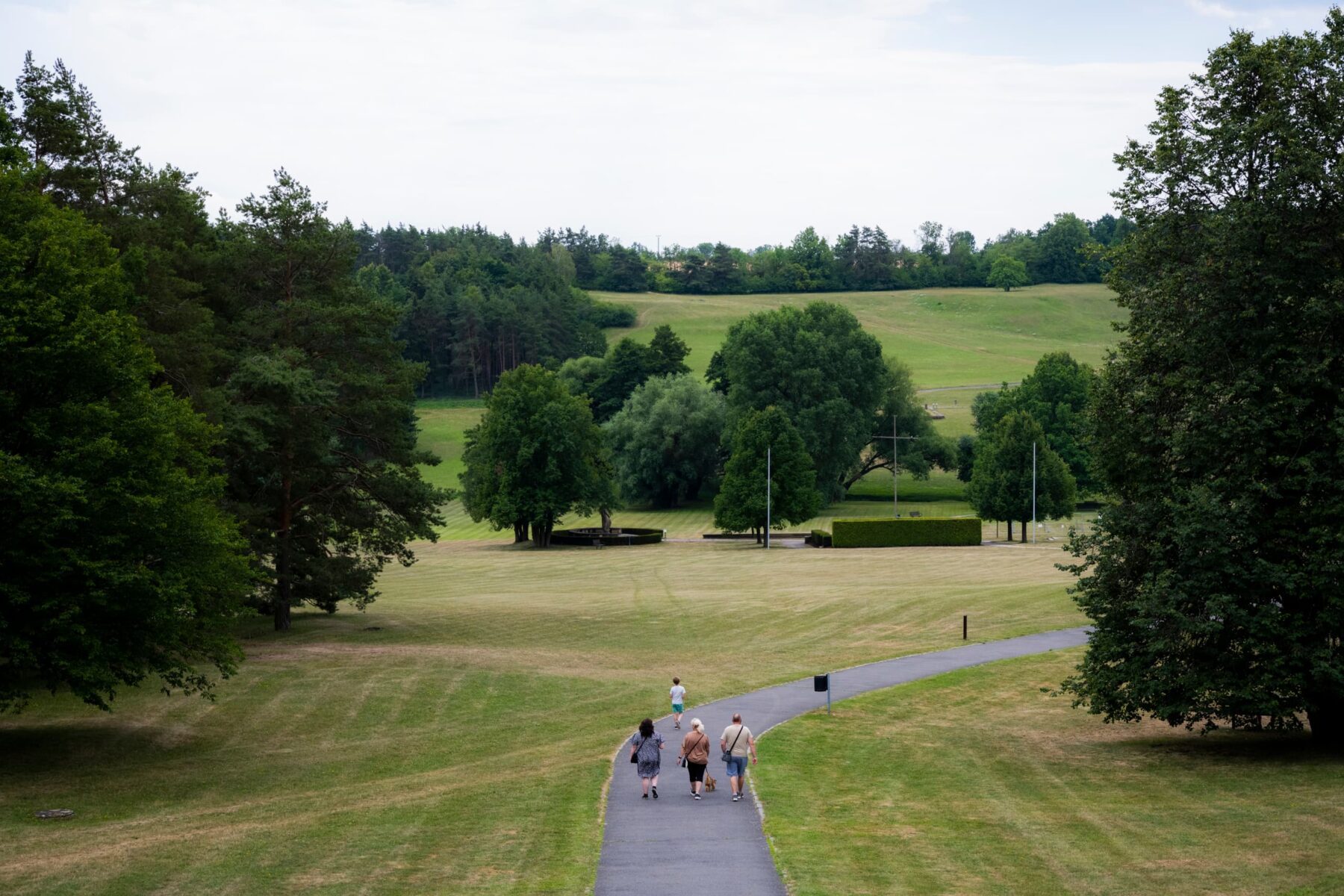 Vier Besucher laufen auf gewundenem Weg durch die weite Parklandschaft der Gedenkstätte Lidice, Bäume und Hügel im Hintergrund.